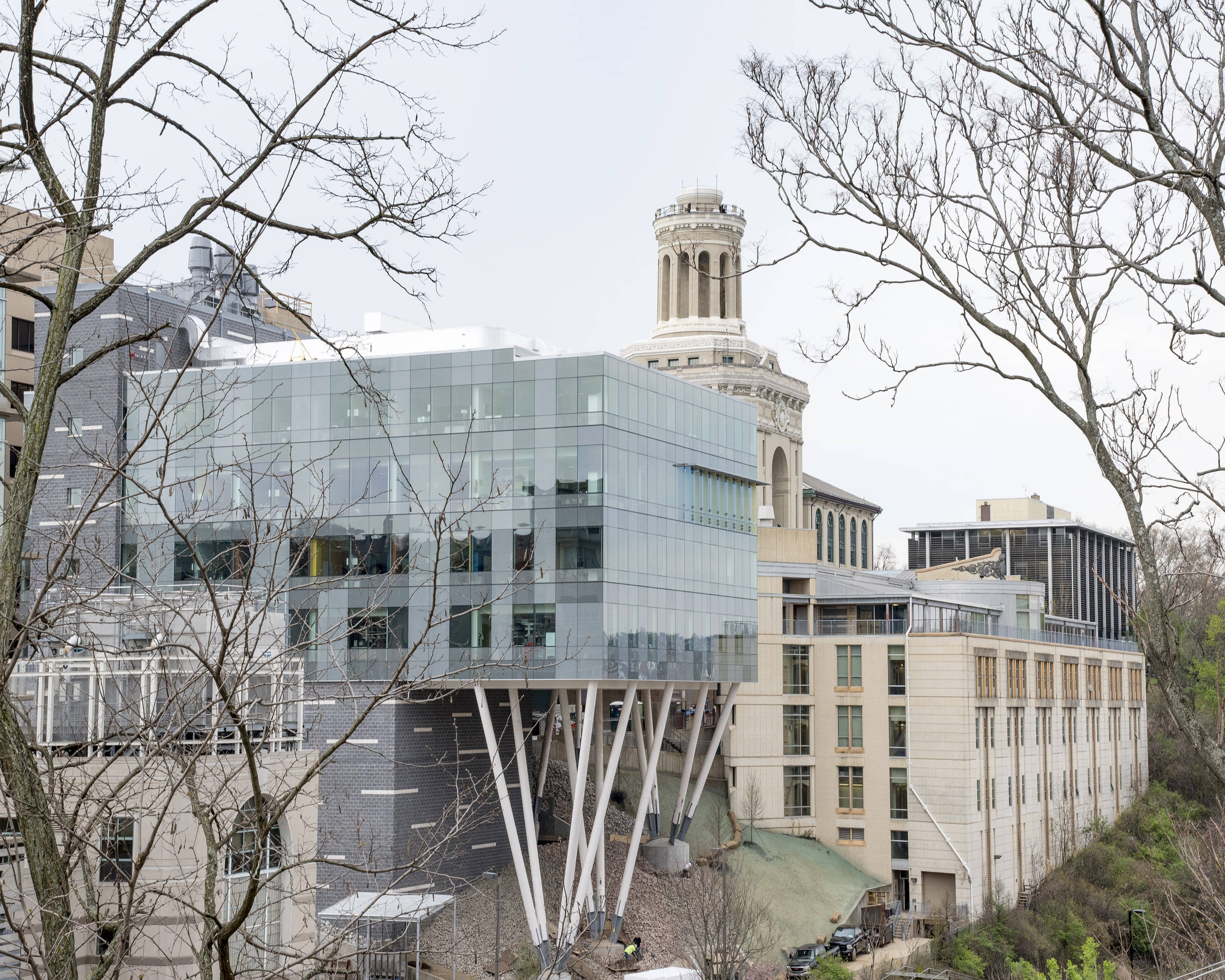Building 桌布 壁紙 — 現代玻璃建築配白色支柱，矗立在歷史石塔旁，由光禿的冬季樹木框出畫面。