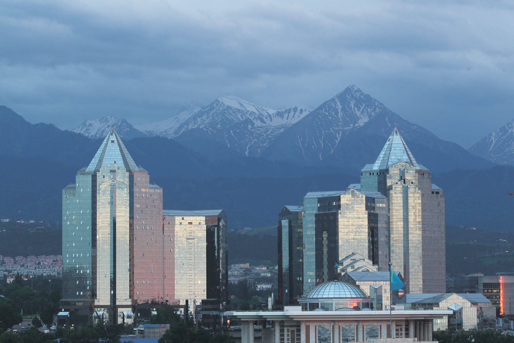 Kazakhstan 桌布 壁紙 — 現代玻璃摩天大樓，屋頂尖銳，以雲天之下積雪覆蓋的山脈為背景。