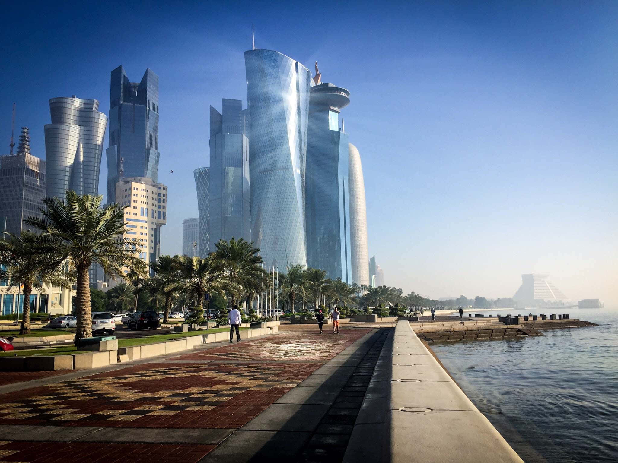 Qatar 桌布 壁紙 — 現代玻璃摩天大樓倒映藍天，旁邊是棕櫚樹成蔭的海濱長廊，有行人漫步。
