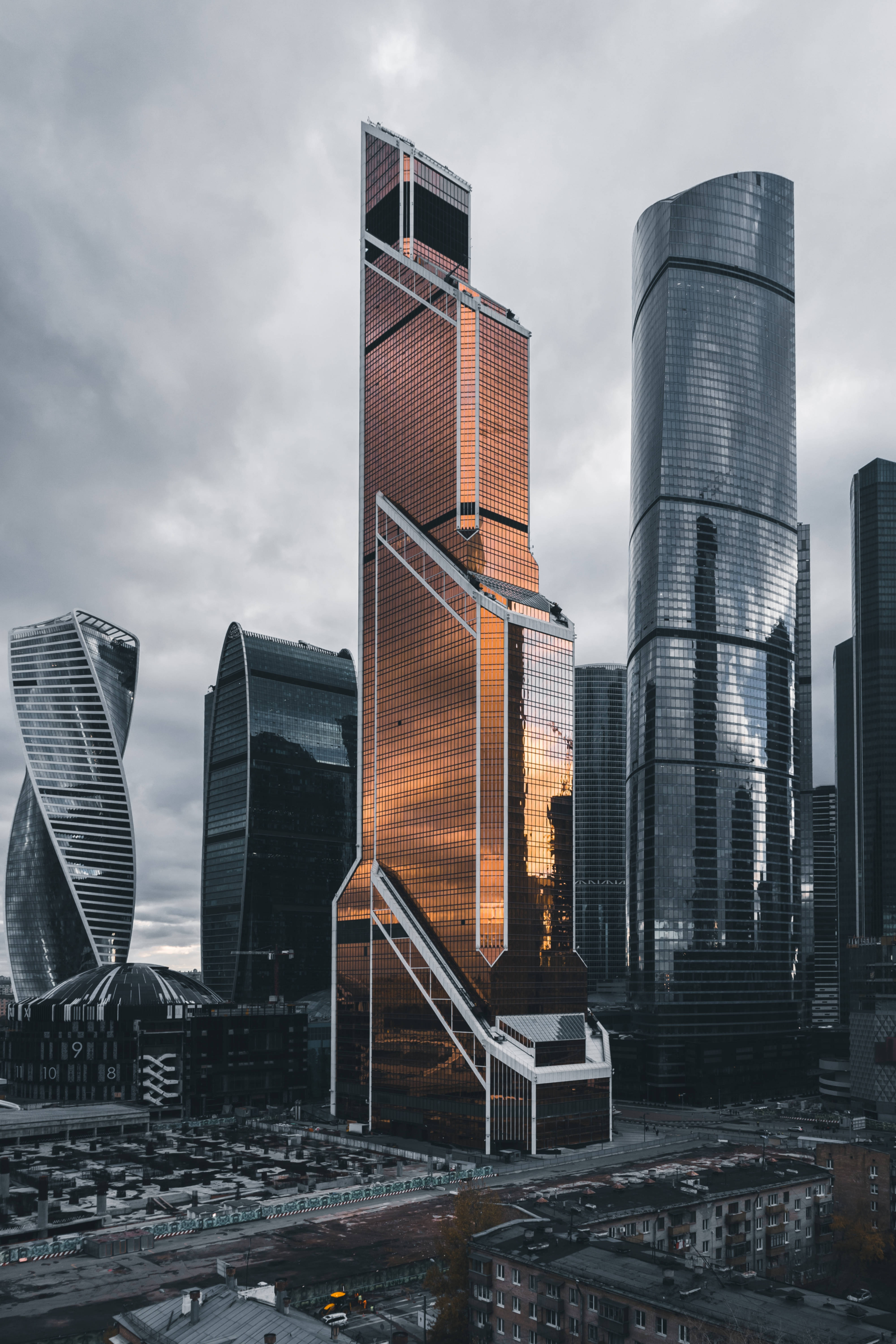 Skyscraper 桌布 壁紙 — 現代玻璃摩天大樓在密集的城市天際線中，映照著金色夕陽色調，背景是陰沉的灰色天空。