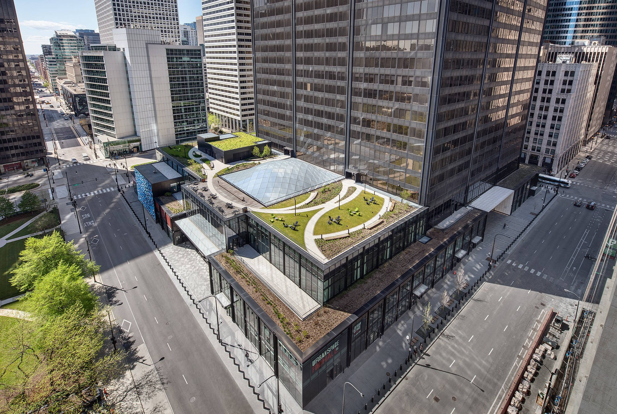 Buildings 桌布 壁紙 — 現代玻璃摩天大樓的鳥瞰圖，擁有茂密的綠色屋頂花園和蜿蜒小徑，周圍是城市街道和其他高樓大廈。