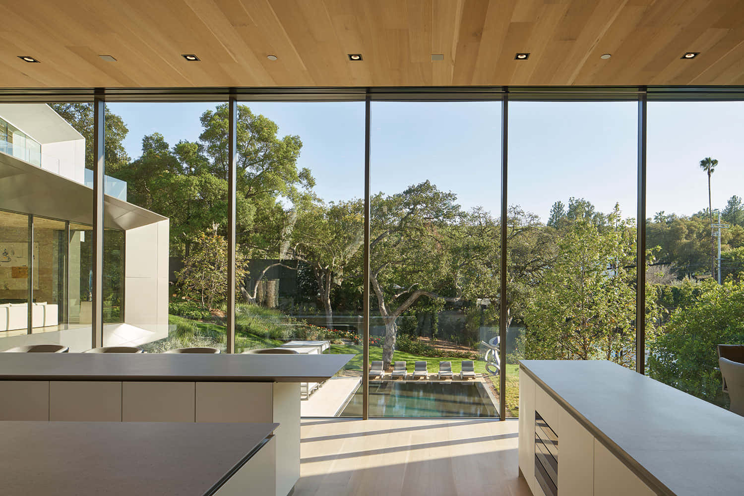 Minimalist Architecture 桌布 壁紙 — 現代玻璃牆內部通向陽光普照的游泳池和茂密的綠樹，在清澈的藍天下。