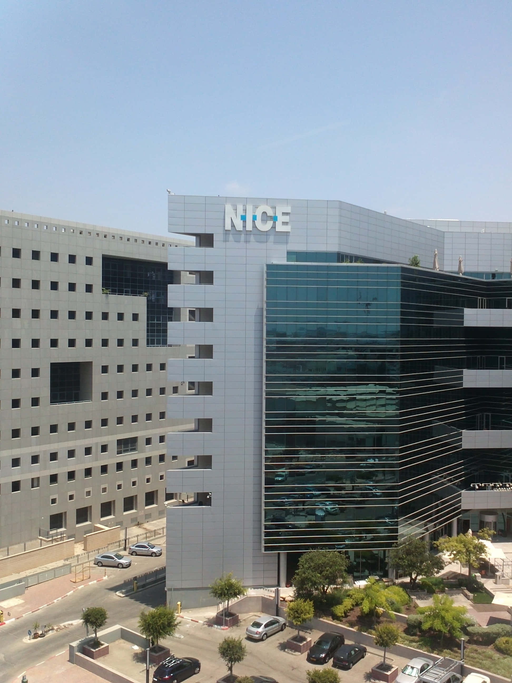 好看 桌布 壁紙 — 現代玻璃鋼筋企業大樓，帶有「NICE」標誌，在晴朗藍天下，下方有停泊的汽車和樹木。