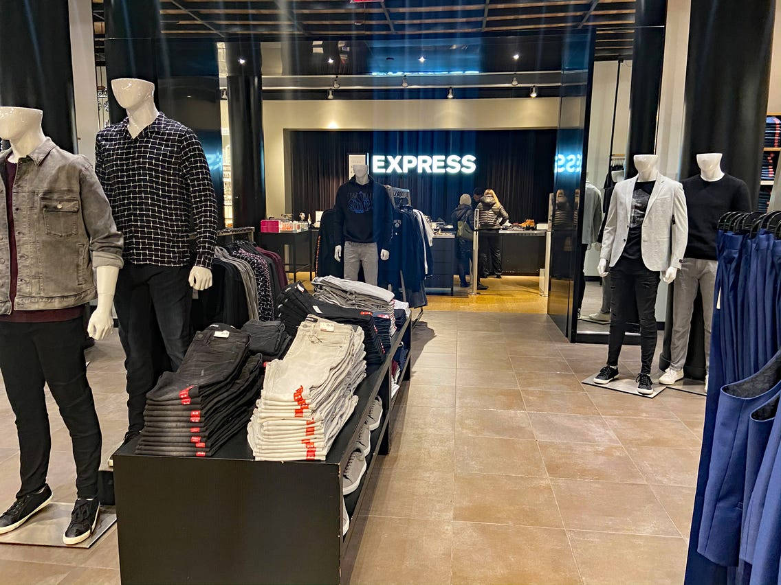 express 桌布 壁紙 — 現代男裝店內部，有模特兒展示休閒服裝，整齊堆疊的牛仔褲在明亮的頭頂燈光下。