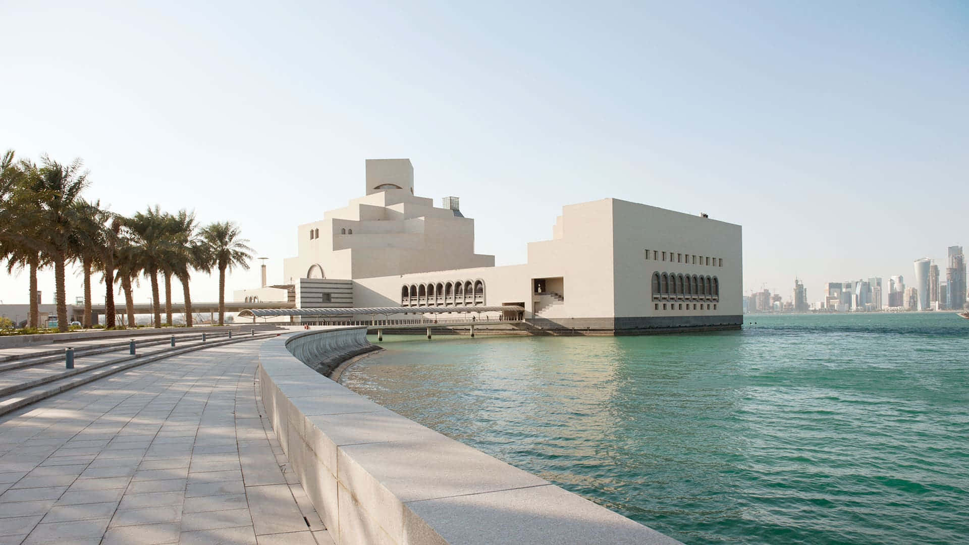 海洋 桌布 壁紙 — 現代白色博物館建築，幾何建築風格，坐落於碧綠水面上，旁邊有棕櫚樹和城市天際線，天空晴朗。