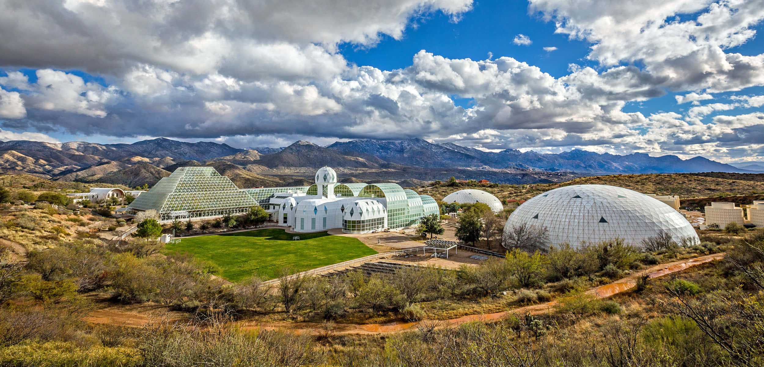 Biosphere 桌布 壁紙 — 現代白色圓頂和玻璃建築，坐落在乾旱沙漠中，周圍有綠色草坪、山脈和戲劇性的多雲天空。