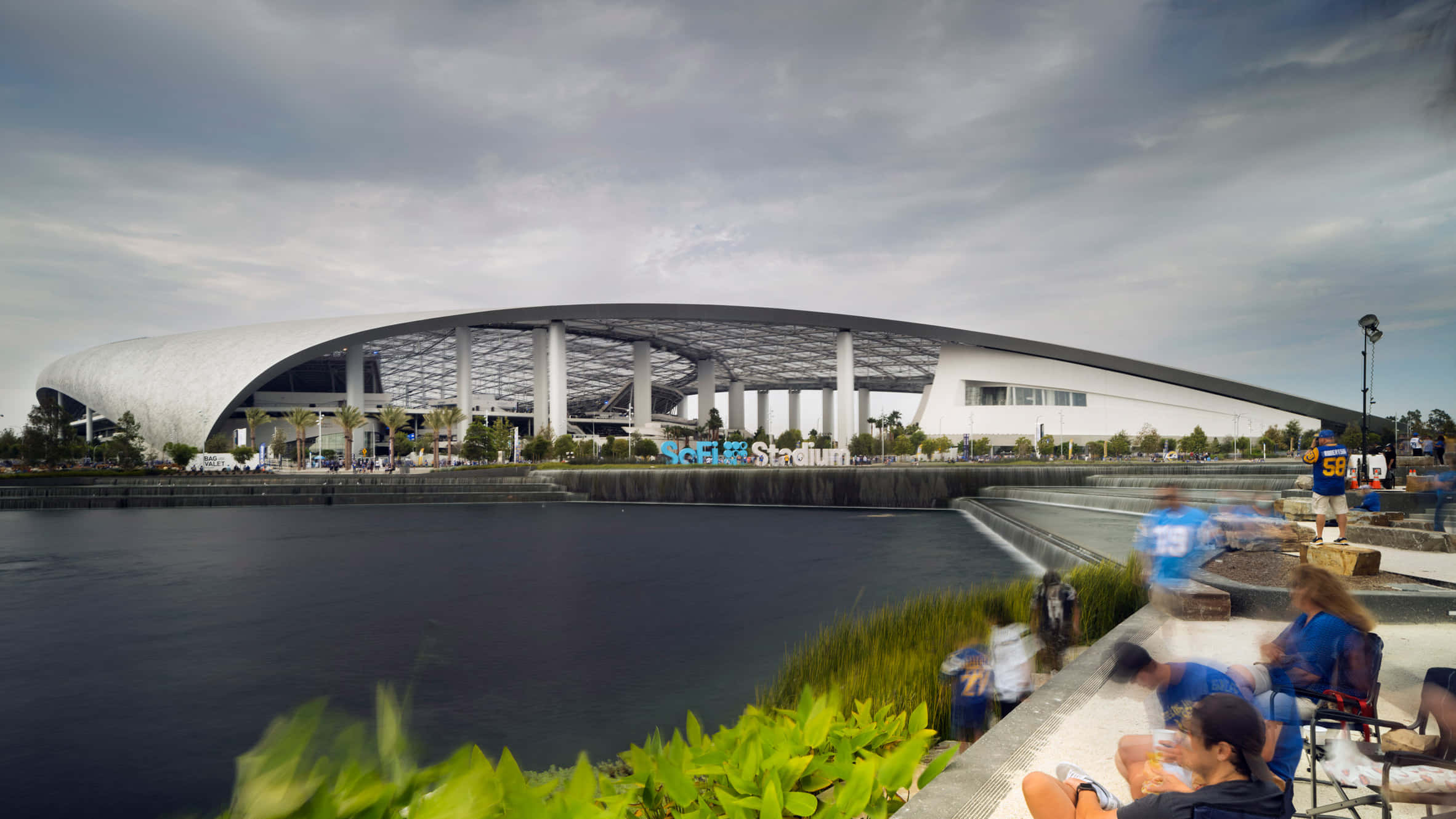 Sofi Stadium 桌布 壁紙 — 現代白色圓頂體育場，帶有「SoFi Stadium」標誌，俯瞰平靜的水面，人們在陰天時在草地上放鬆。
