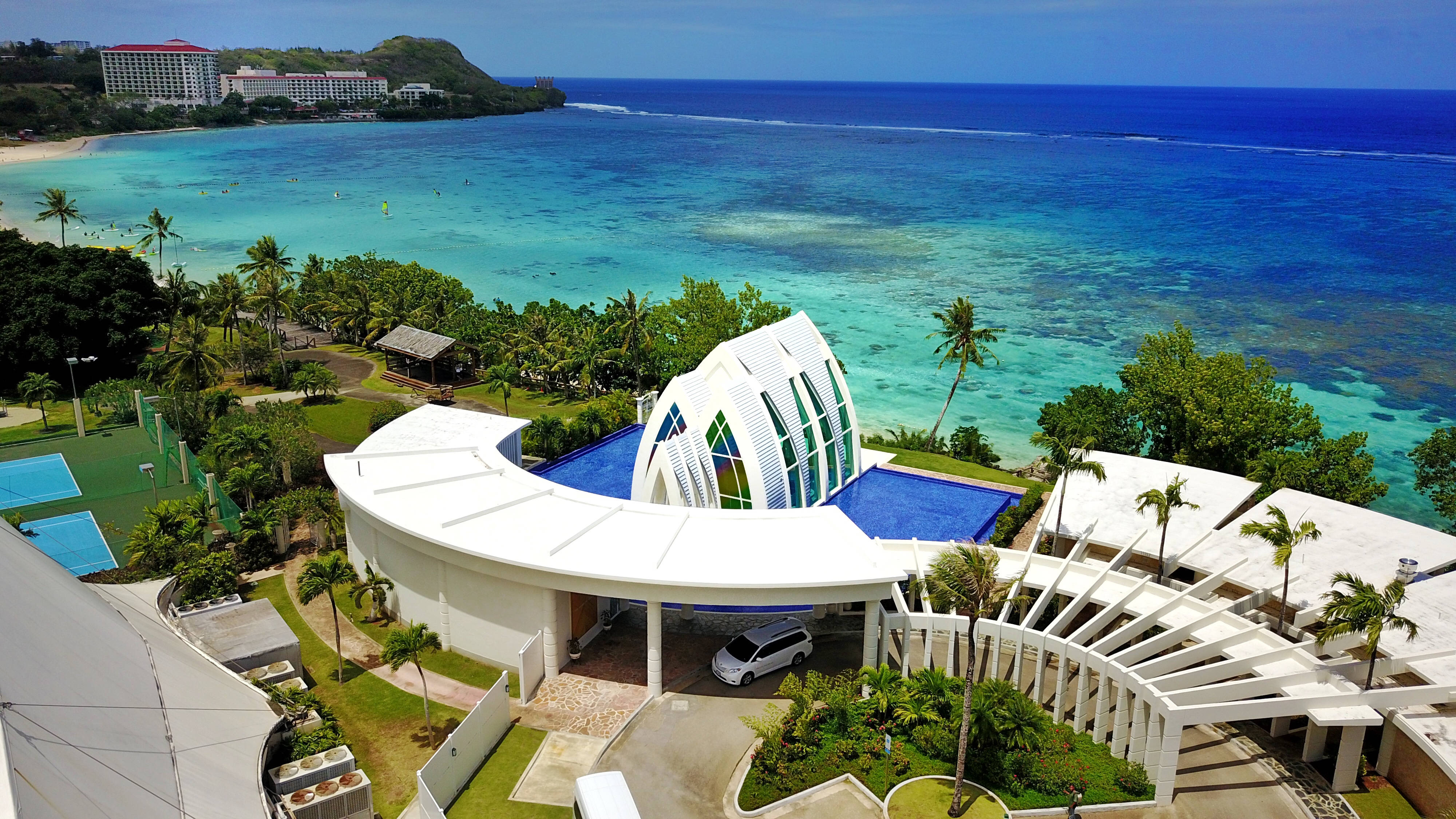 Guam 桌布 壁紙 — 現代白色度假村的鳥瞰圖，設有弧形玻璃教堂，俯瞰碧綠海洋和棕櫚樹。