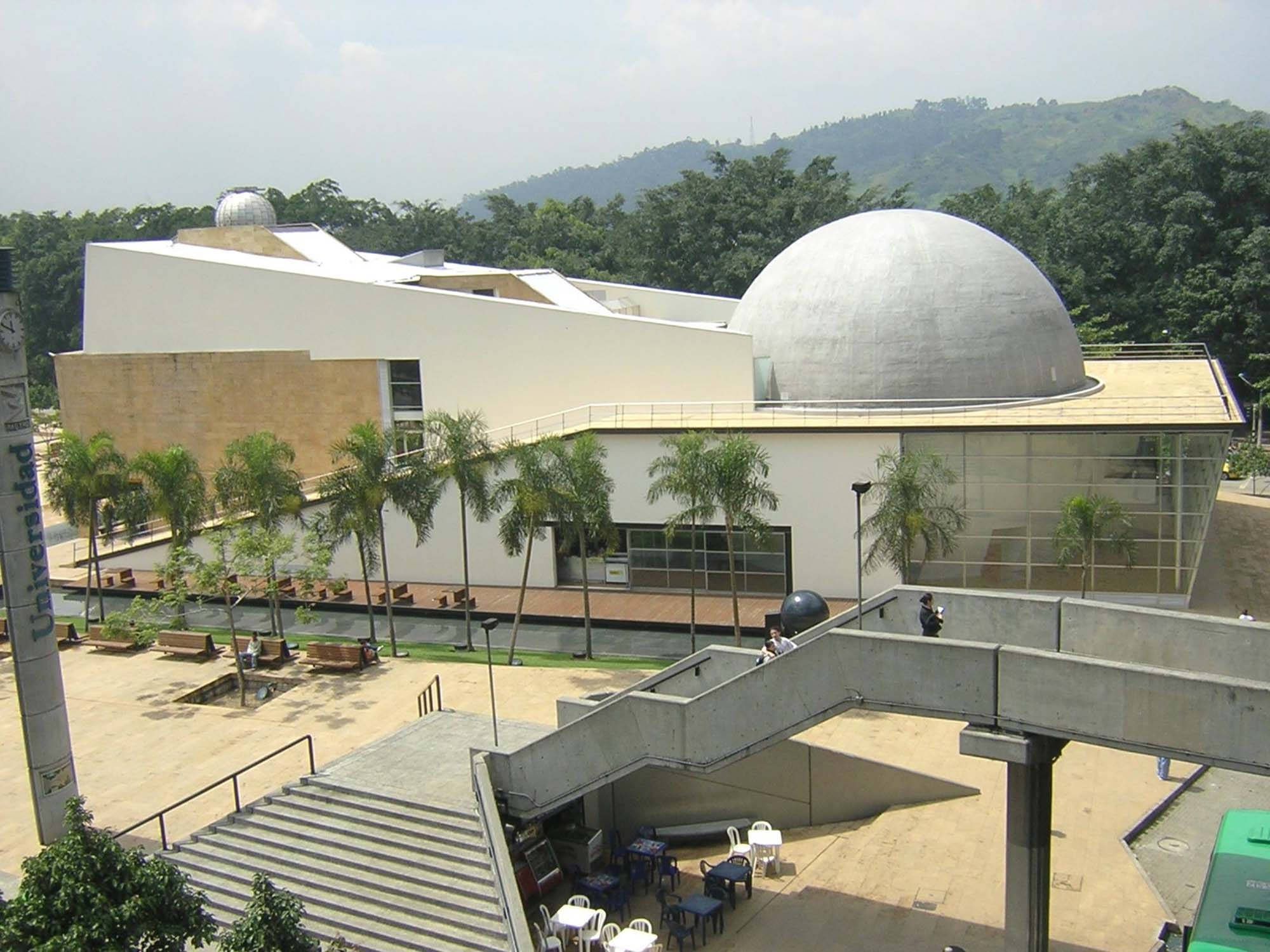 Medellin 桌布 壁紙 — 現代白色建築，擁有大型銀色圓頂和棕櫚樹，以綠色山丘為背景，天空明亮。