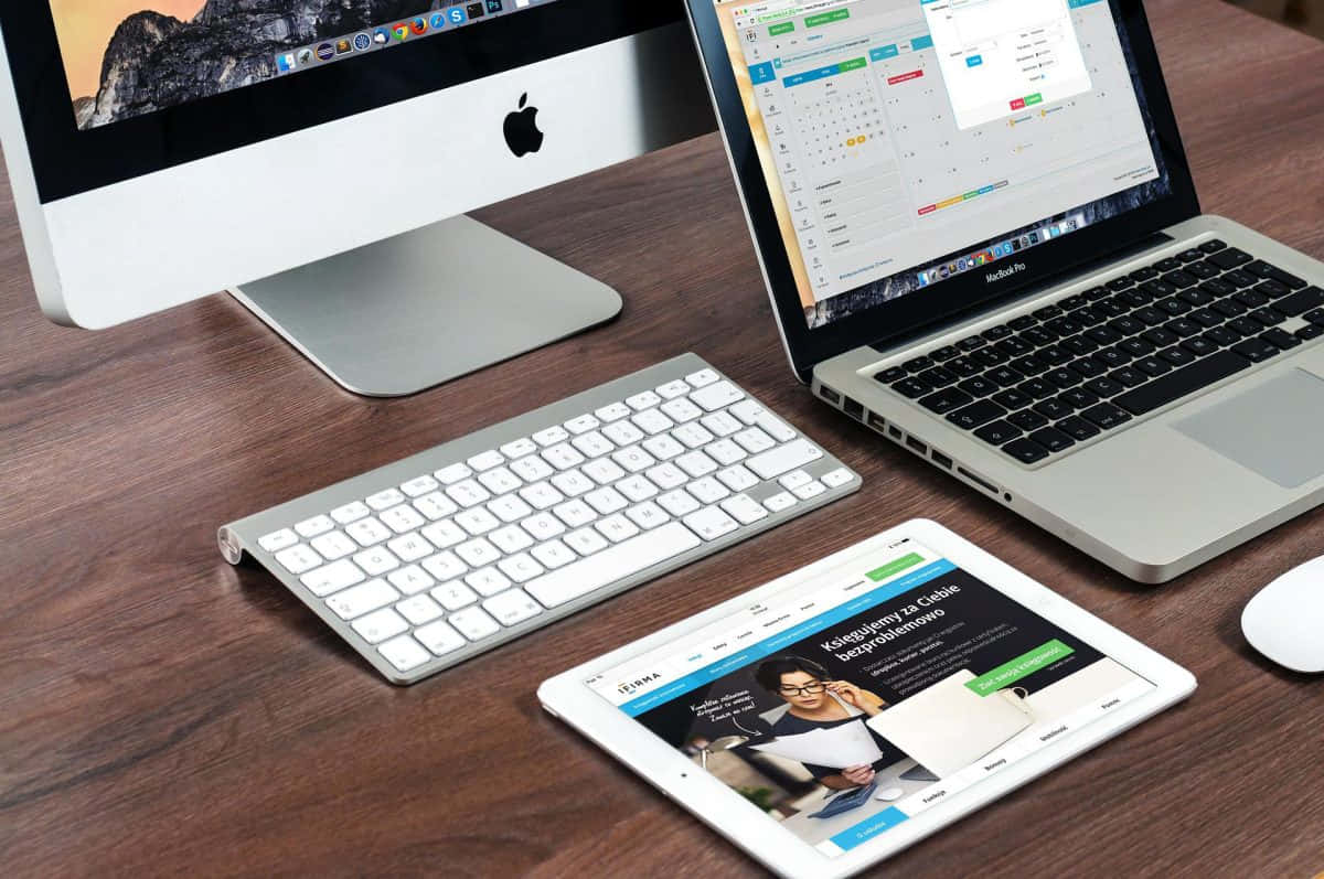 Mac 桌布 壁紙 — 現代科技設置，配備 iMac、筆記型電腦、平板電腦和鍵盤，置於木桌上，顯示生產力應用程式。