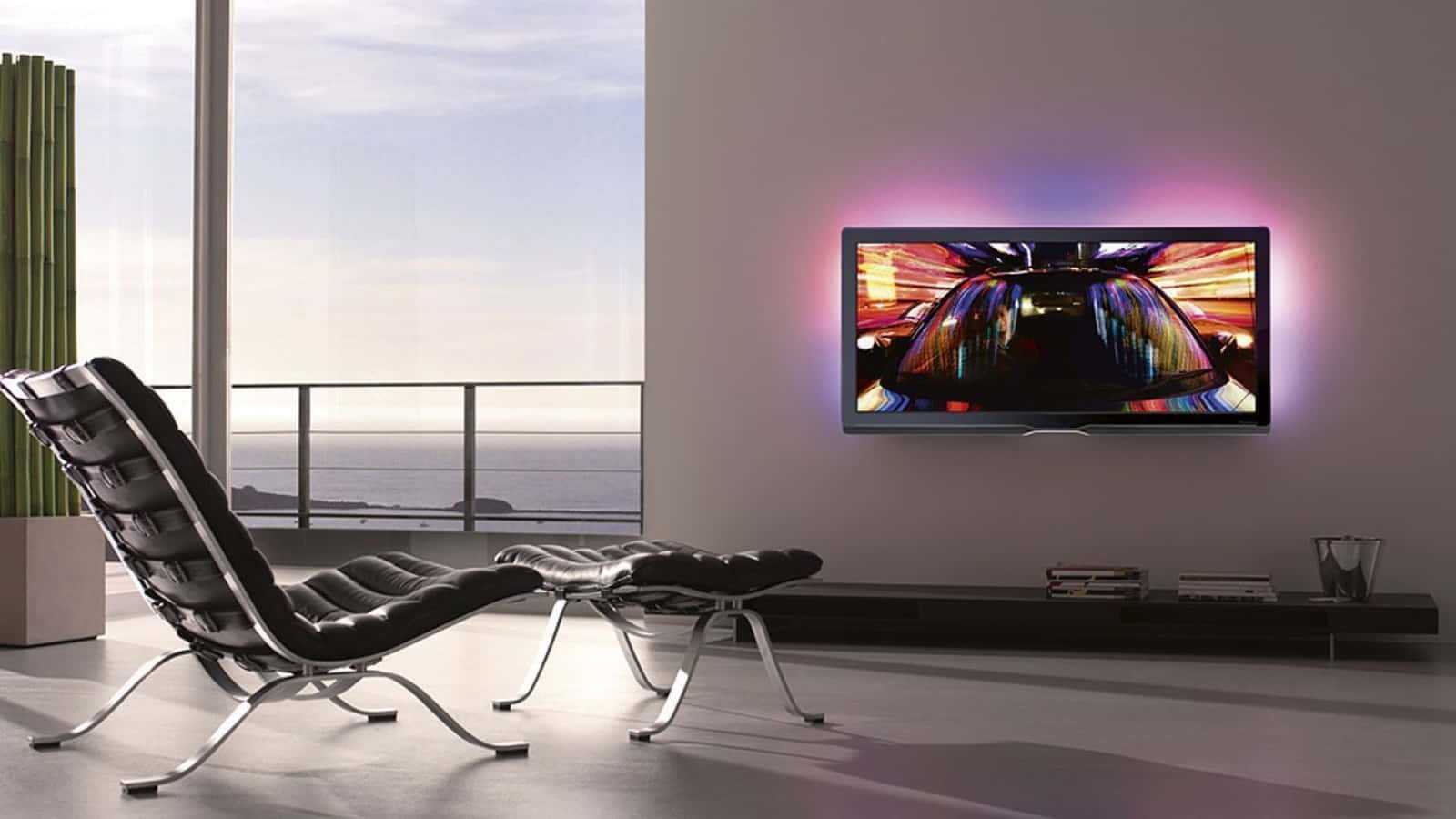 tv 桌布 壁紙 — 現代簡約客廳，黑色休閒椅面向電視，電視播放著色彩鮮豔的賽車場景，背景是明亮的海景。