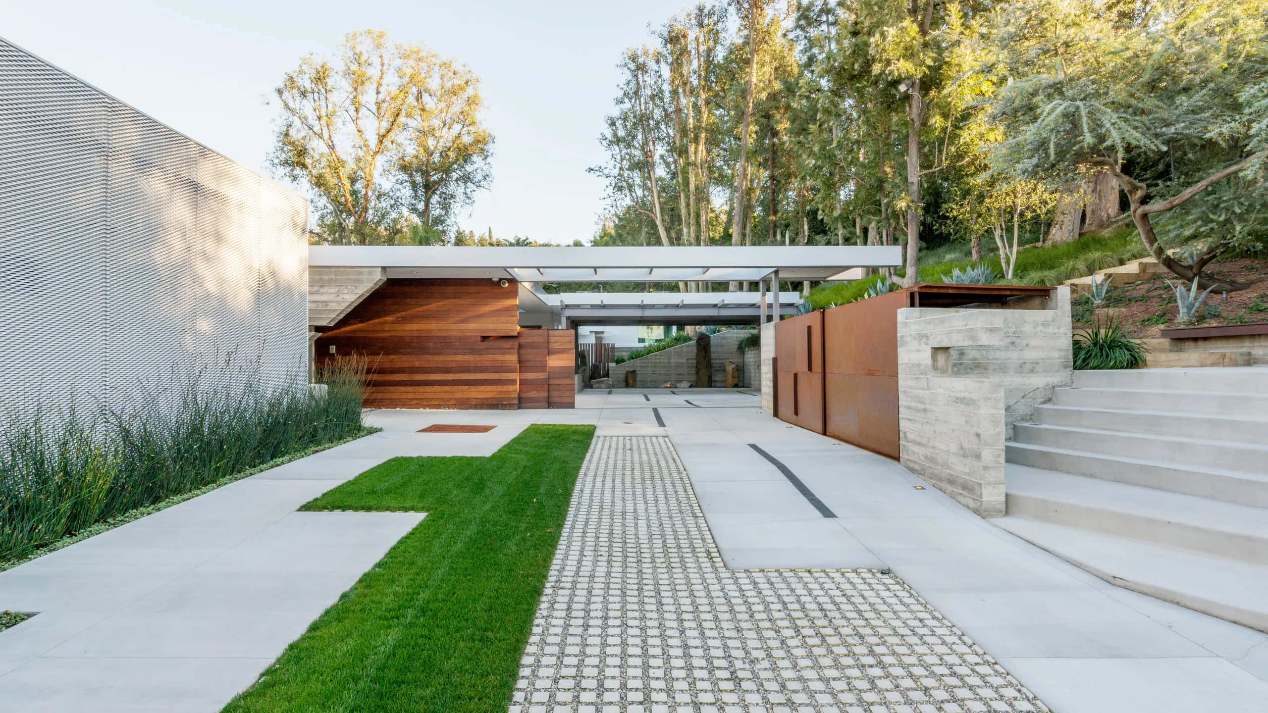 Driveway Ideas 桌布 壁紙 — 現代簡約家居入口，設有混凝土小徑、草地點綴和鏽蝕鋼牆，周圍環繞著高大的樹木，在明亮的日光下。