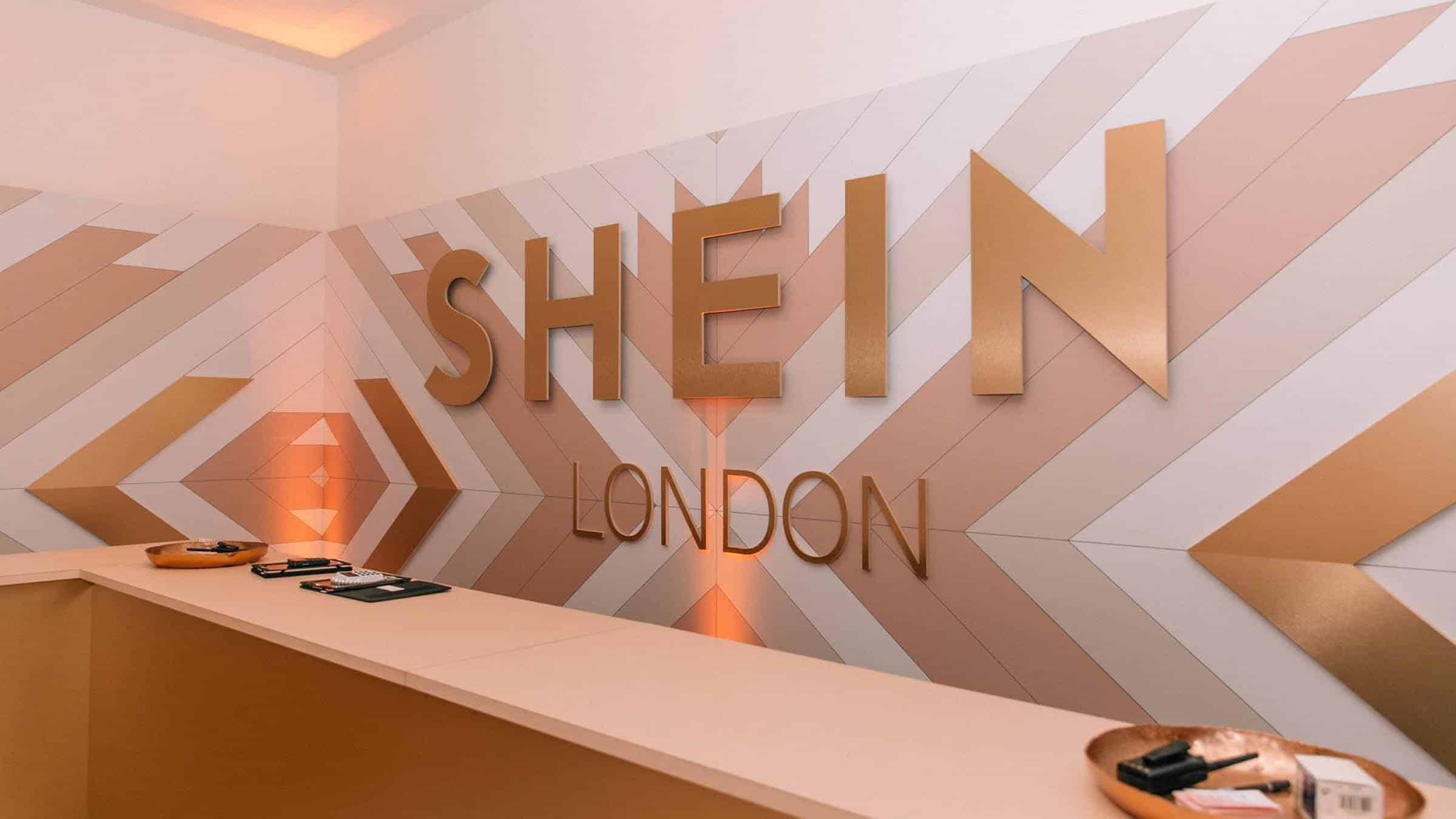 Shein 桌布 壁紙 — 現代簡約接待區，金色 'SHEIN LONDON' 標誌，背景為幾何人字形牆壁，採用溫暖的桃色和白色色調。