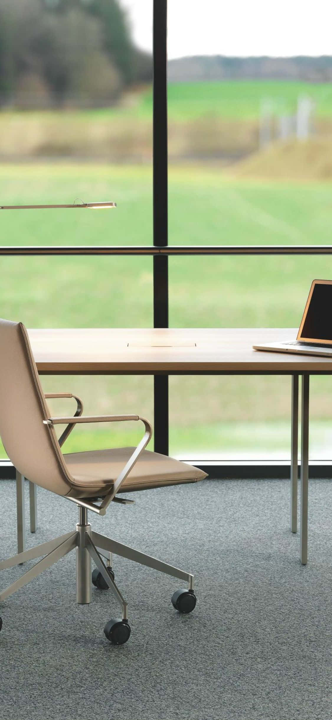 iPhone Xs Desk 桌布 壁紙 — 現代簡約辦公室書桌，配有棕褐色椅子和筆記型電腦，位於大窗旁，俯瞰綠色田野。