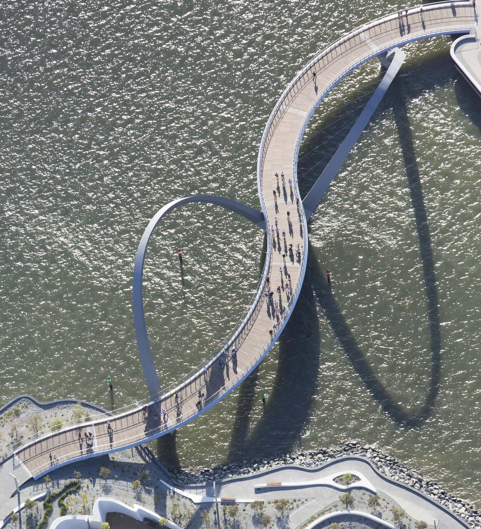 bridge 桌布 壁紙 — 現代蜿蜒行人天橋的鳥瞰圖，在漣漪的水面上投下長長的陰影，人們沿著其彎曲的路徑行走。