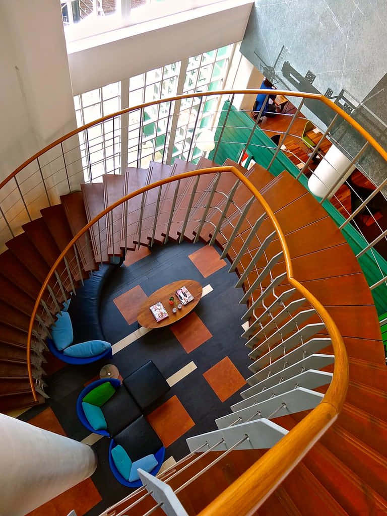 Interior Design 桌布 壁紙 — 現代螺旋樓梯的鳥瞰圖，樓梯採用木質踏板和金屬欄杆，環繞著一個中央座位區，擺放著色彩繽紛的椅子。