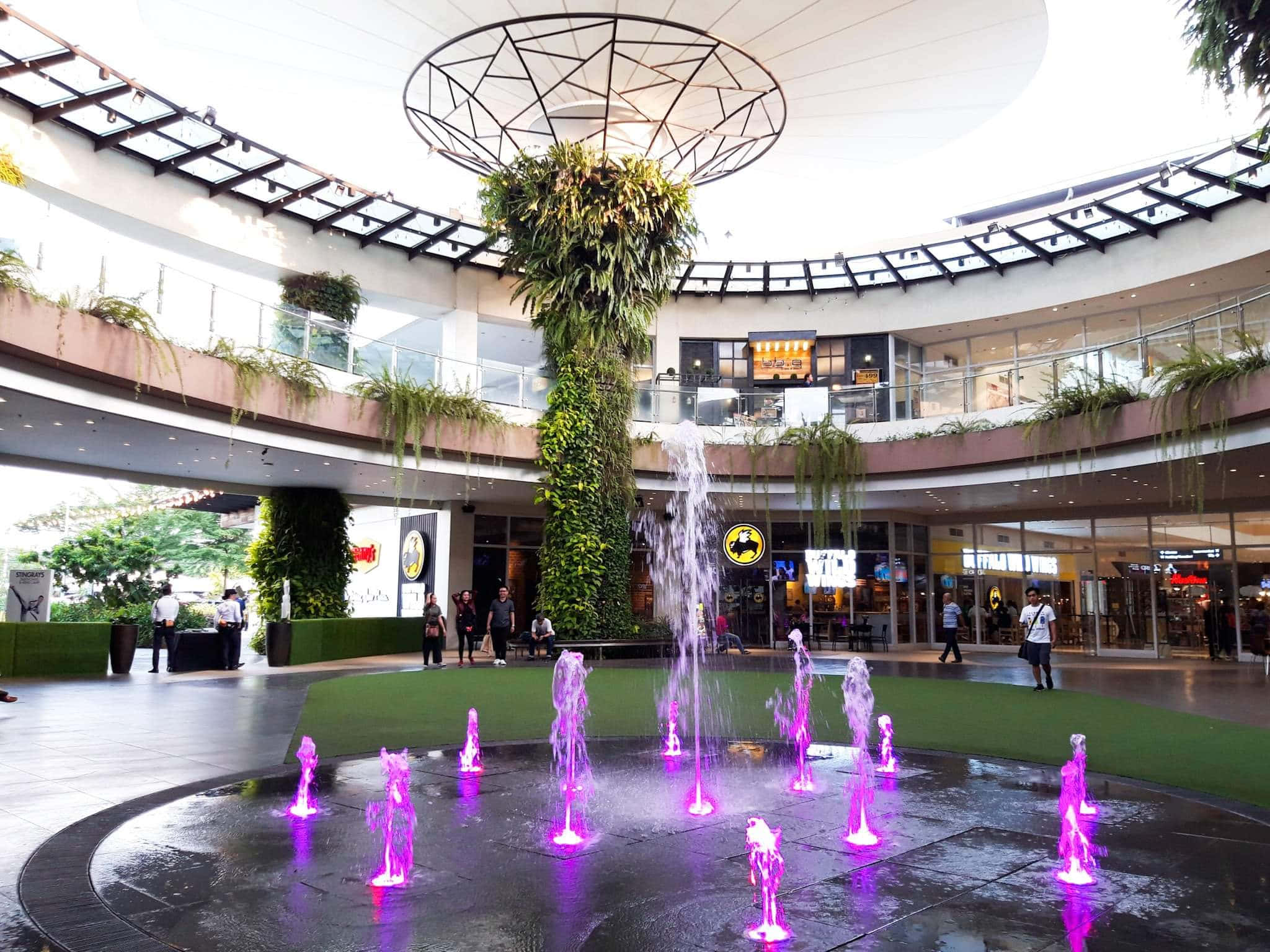 Mall 桌布 壁紙 — 現代購物中心中庭設有中央噴泉，散發著紫色光芒，周圍環繞著茂密的綠植和多層步道。