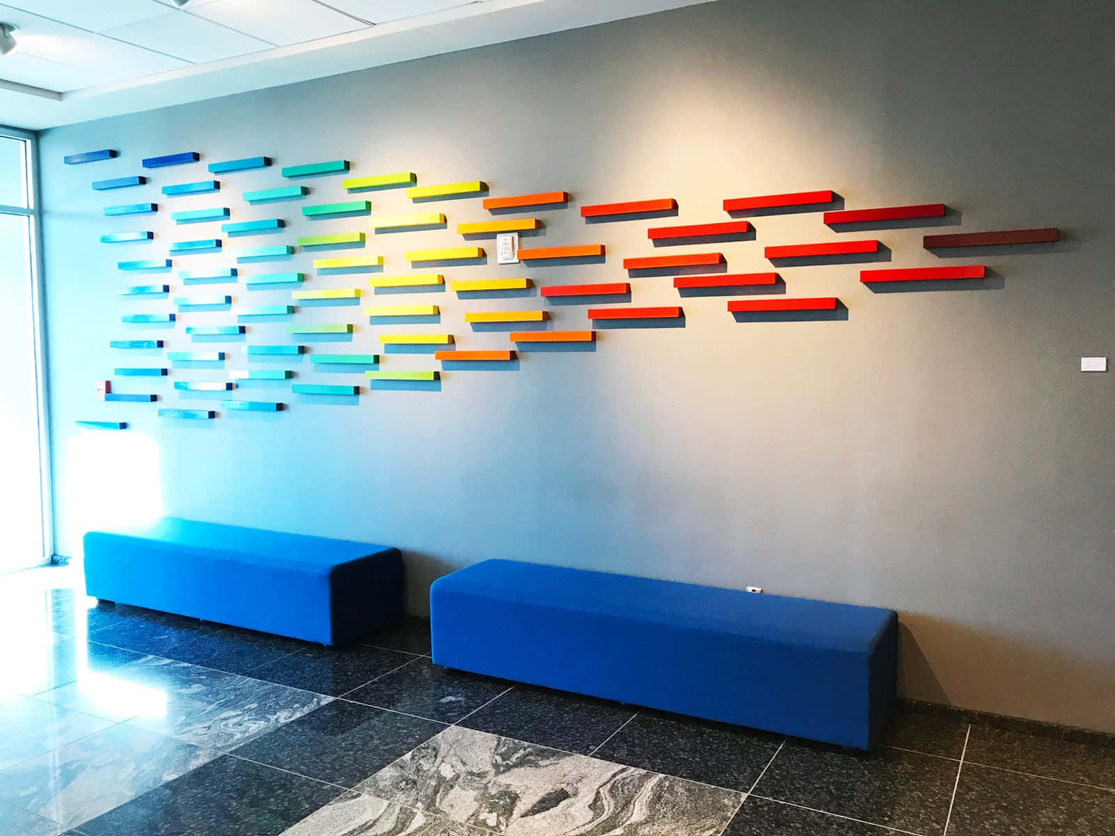 Installation Art 桌布 壁紙 — 現代辦公室大廳，灰色牆壁上兩個藍色長凳上方有鮮豔彩虹色浮動層架藝術品。