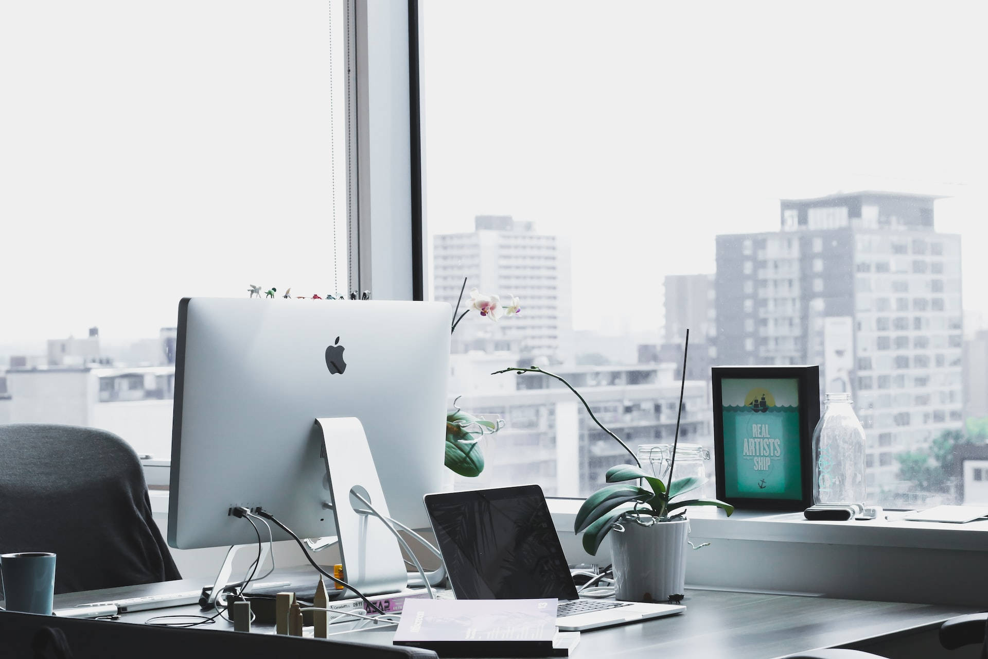 Office Space 桌布 壁紙 — 現代辦公室書桌，配備 iMac 筆記型電腦和蘭花植物，透過大窗戶俯瞰城市天際線。