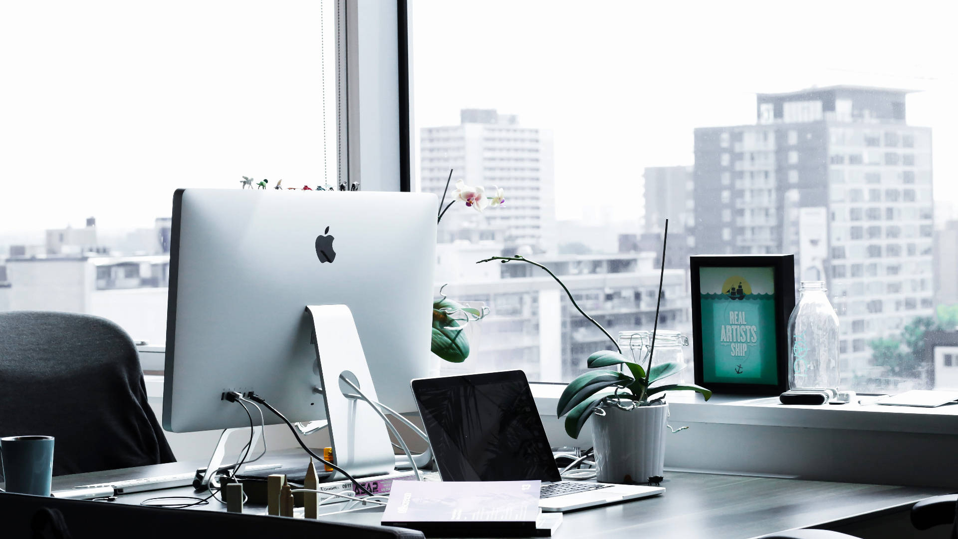 電腦 桌布 壁紙 — 現代辦公室書桌，配備 iMac 和筆記型電腦，位於窗邊，俯瞰城市天際線，並有盆栽植物和裱框藝術品。