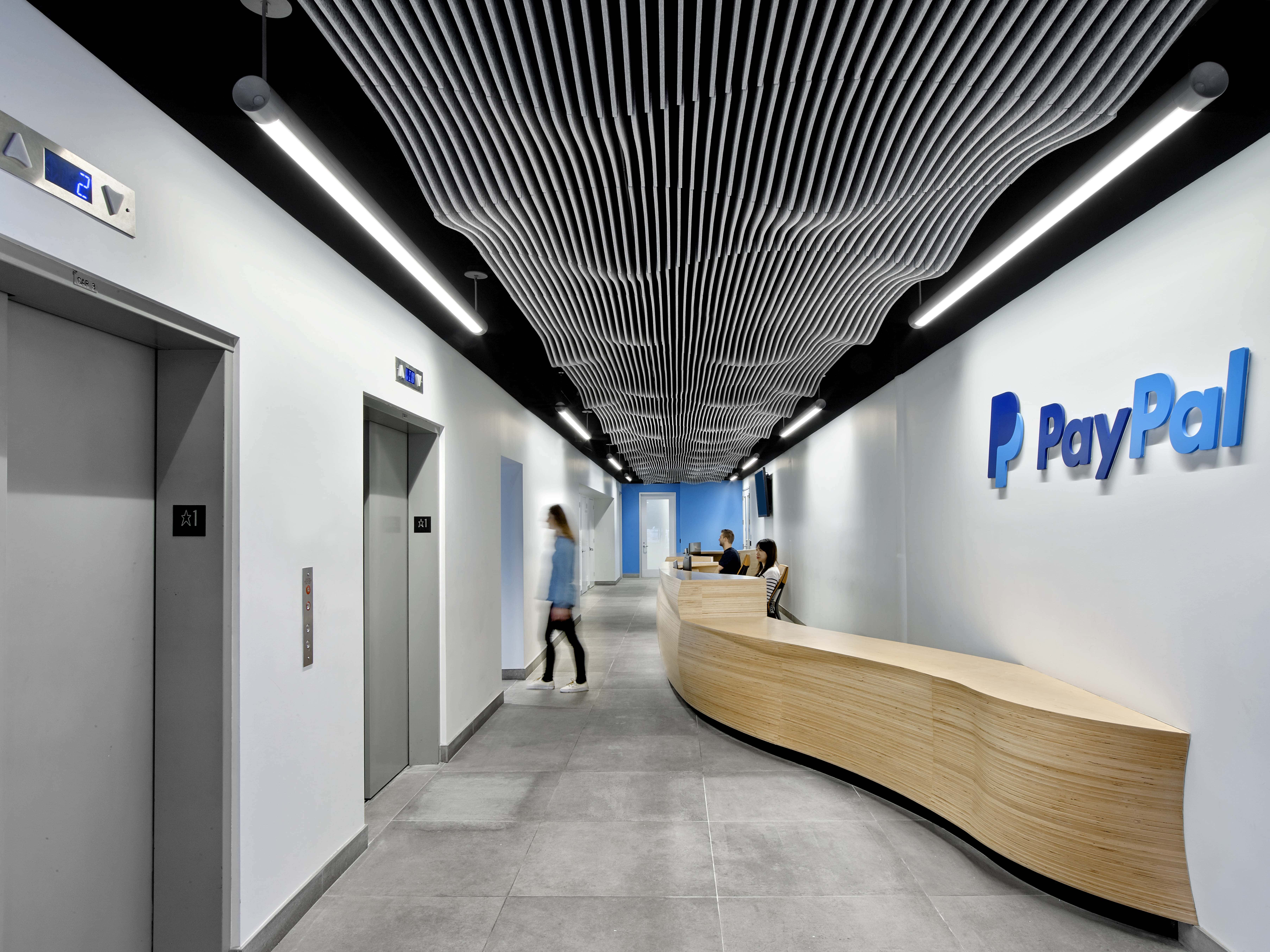 PayPal 桌布 壁紙 — 現代辦公室走廊，設有弧形木製接待桌和波浪形天花板，在明亮燈光下。