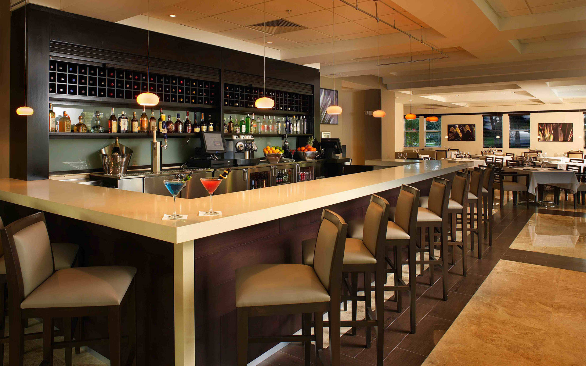 Bar 桌布 壁紙 — 現代酒店酒吧，設有拋光木質吧檯、兩杯色彩繽紛的雞尾酒、兩張吧凳，以及暖色吊燈下擺滿酒瓶的層架。