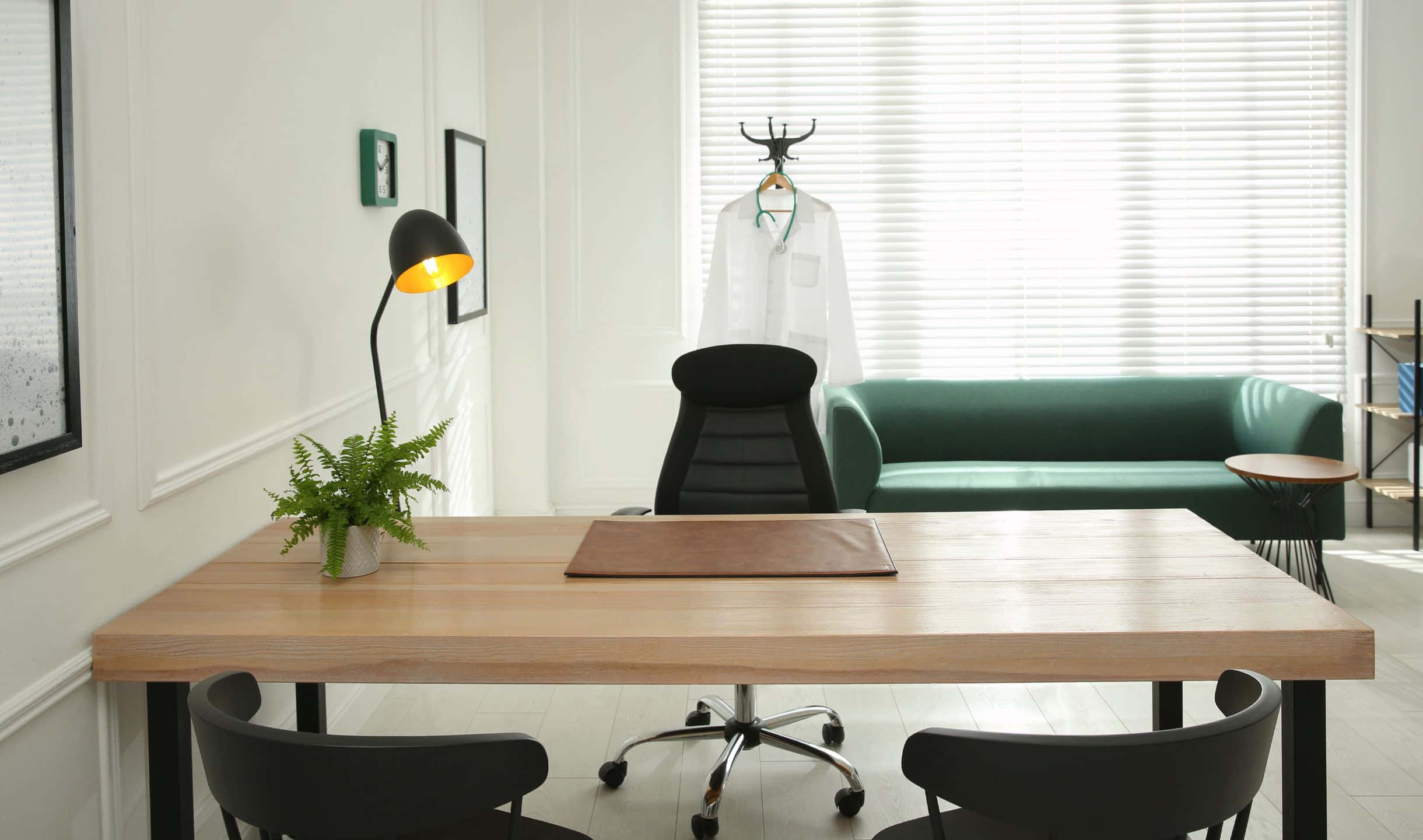 Office 桌布 壁紙 — 現代醫生診所，配有木桌、黑色椅子、綠色沙發和掛在窗邊的白袍。
