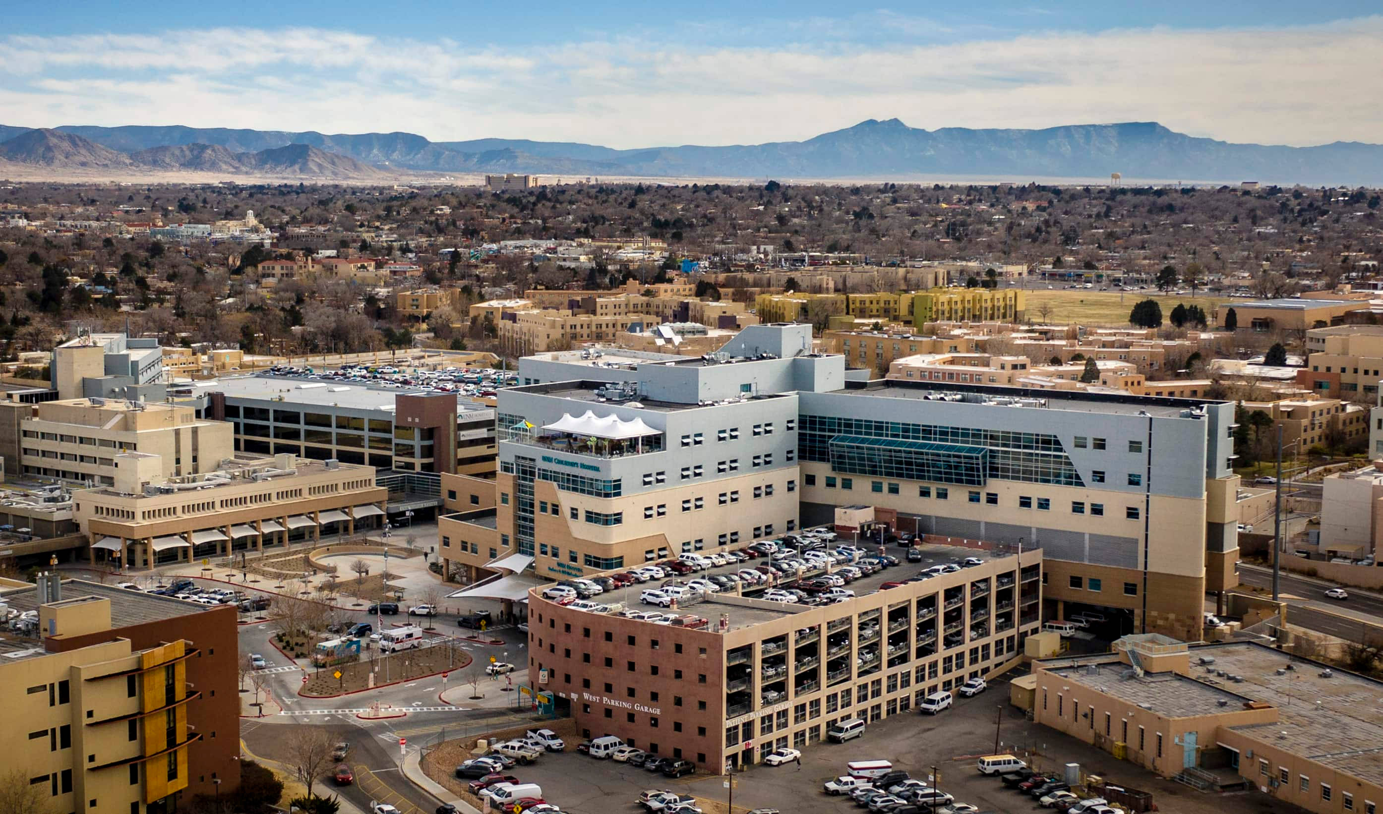 Hospital 桌布 壁紙 — 現代醫院建築群的鳥瞰圖，設有停車場結構和周圍的城市擴張，背景是遠處的山脈，天空部分多雲。