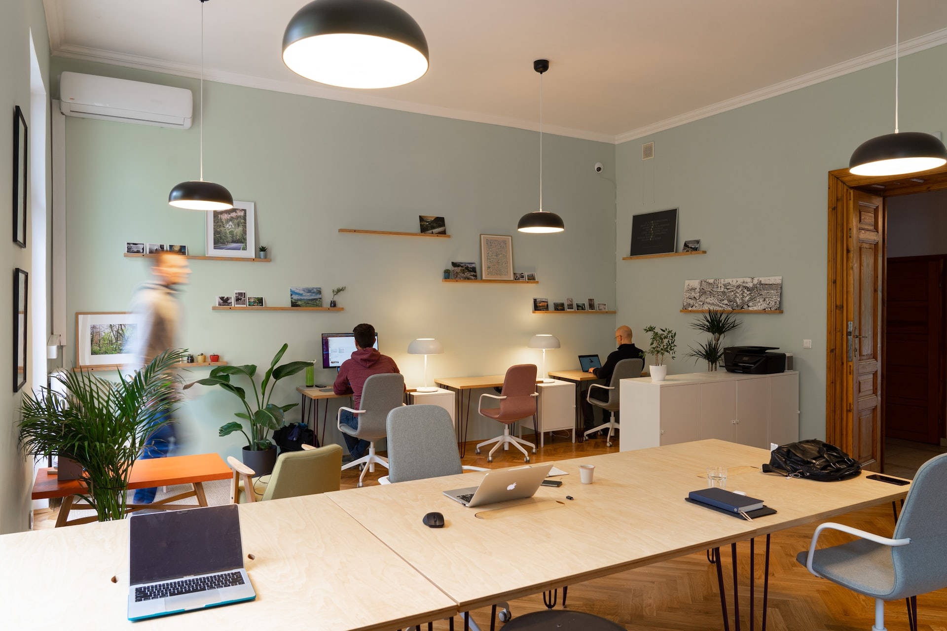 Office Space 桌布 壁紙 — 現代開放式辦公室，配有淺色木桌、筆記型電腦、吊燈，人們在平靜協作的空間中工作。