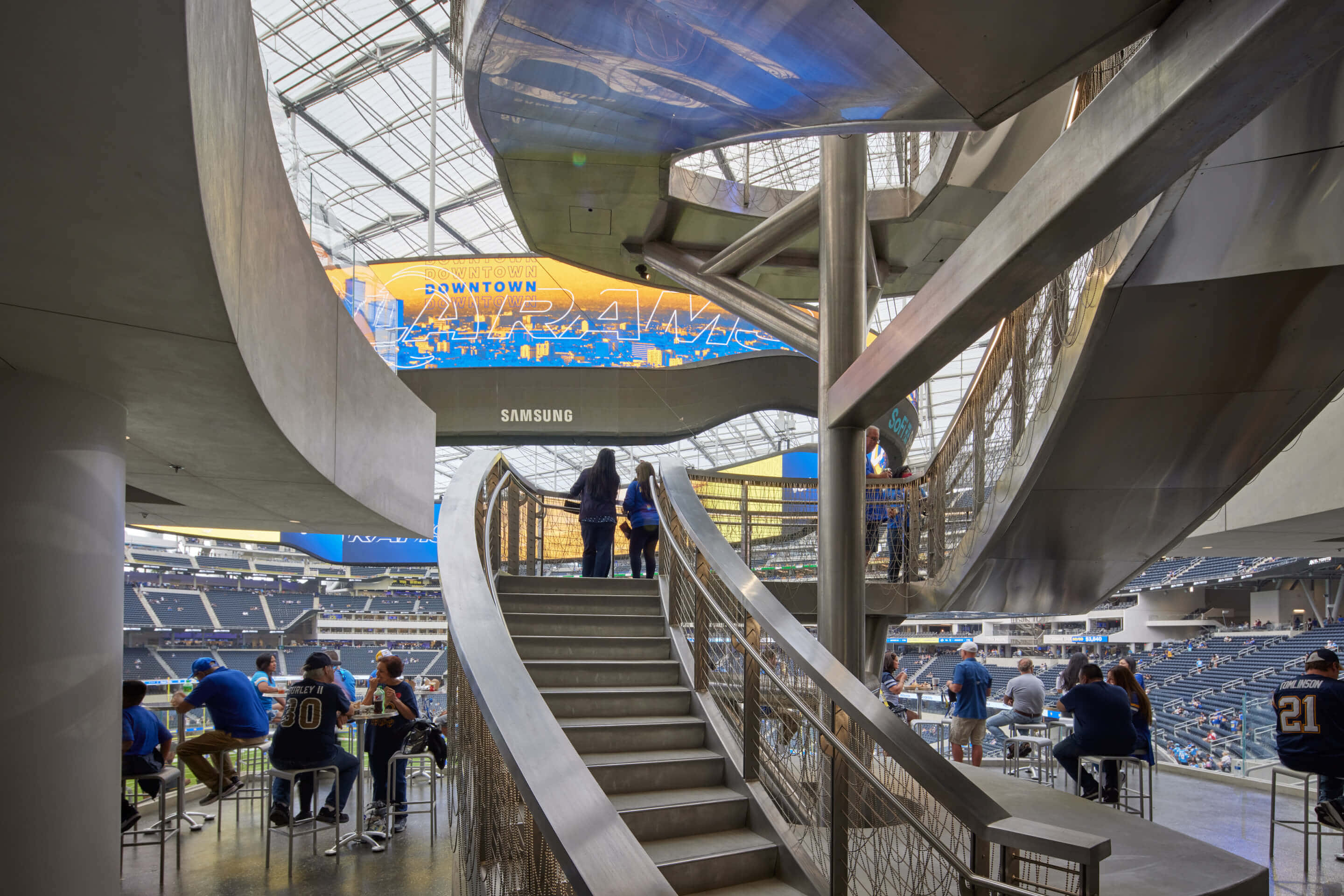 Sofi Stadium 桌布 壁紙 — 現代體育館內部，設有彎曲的金屬樓梯，身穿藍色球衣的球迷在大型場館享受比賽日。