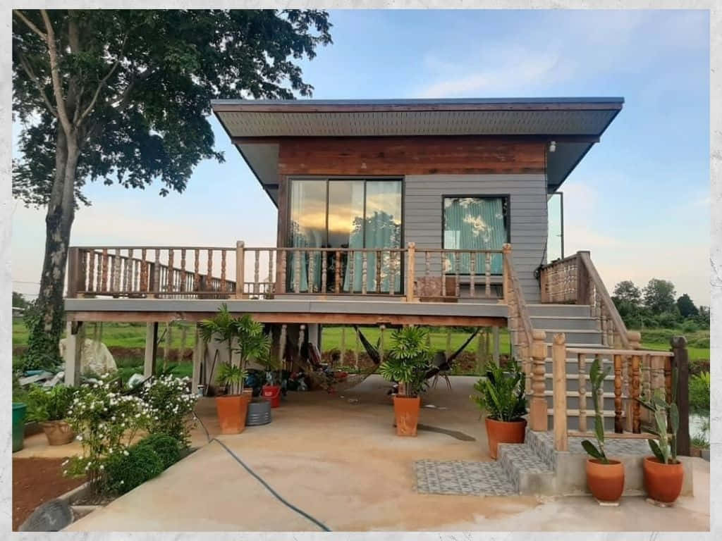 cabin 桌布 壁紙 — 現代高架木屋，配有玻璃窗和陽台，周圍環繞著茂密的綠植和盆栽植物，在清澈的藍天下。
