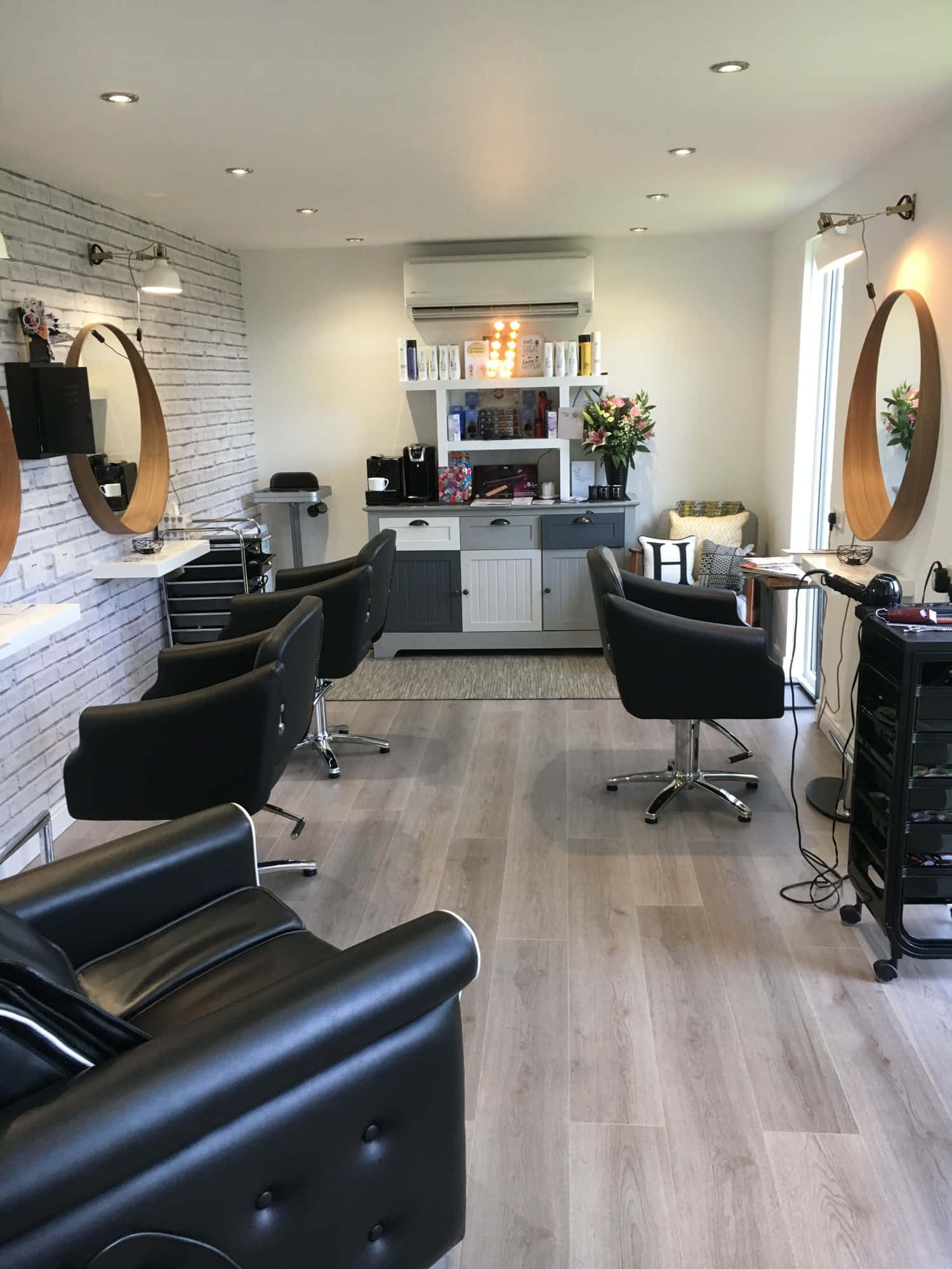 Hair Salon 桌布 壁紙 — 現代髮廊，配備黑色皮革椅、木地板和白色磚牆，沐浴在自然光中。