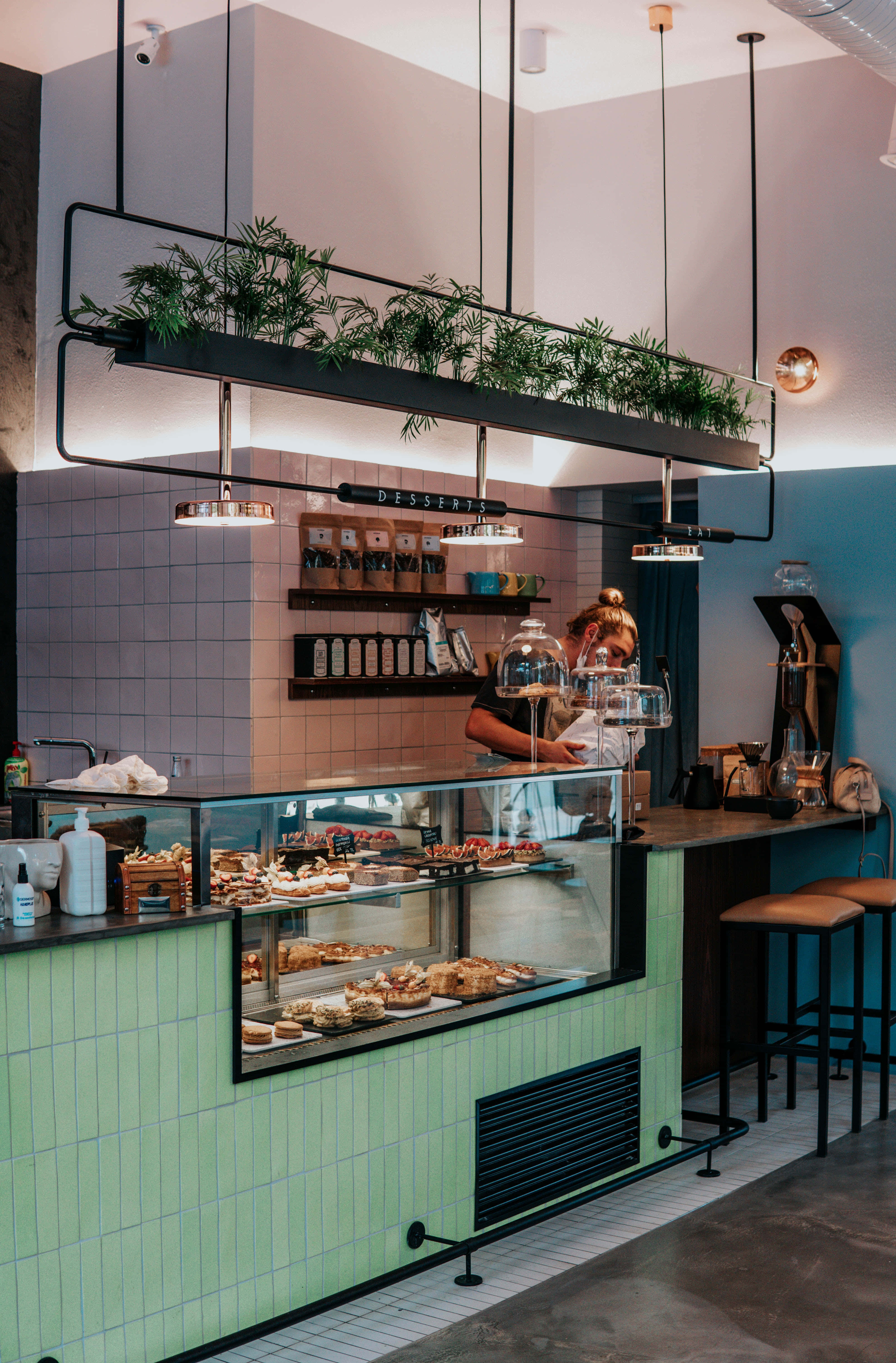 Bakery 桌布 壁紙 — 現代麵包店內部，薄荷綠色瓷磚櫃檯展示玻璃下的糕點，員工在櫃檯後工作。
