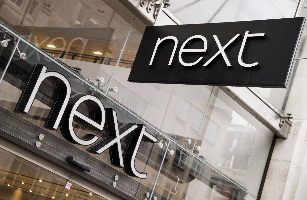 Store 桌布 壁紙 — 現代黑白「next」商店標誌，以粗體字安裝在玻璃外牆上，反映城市環境。