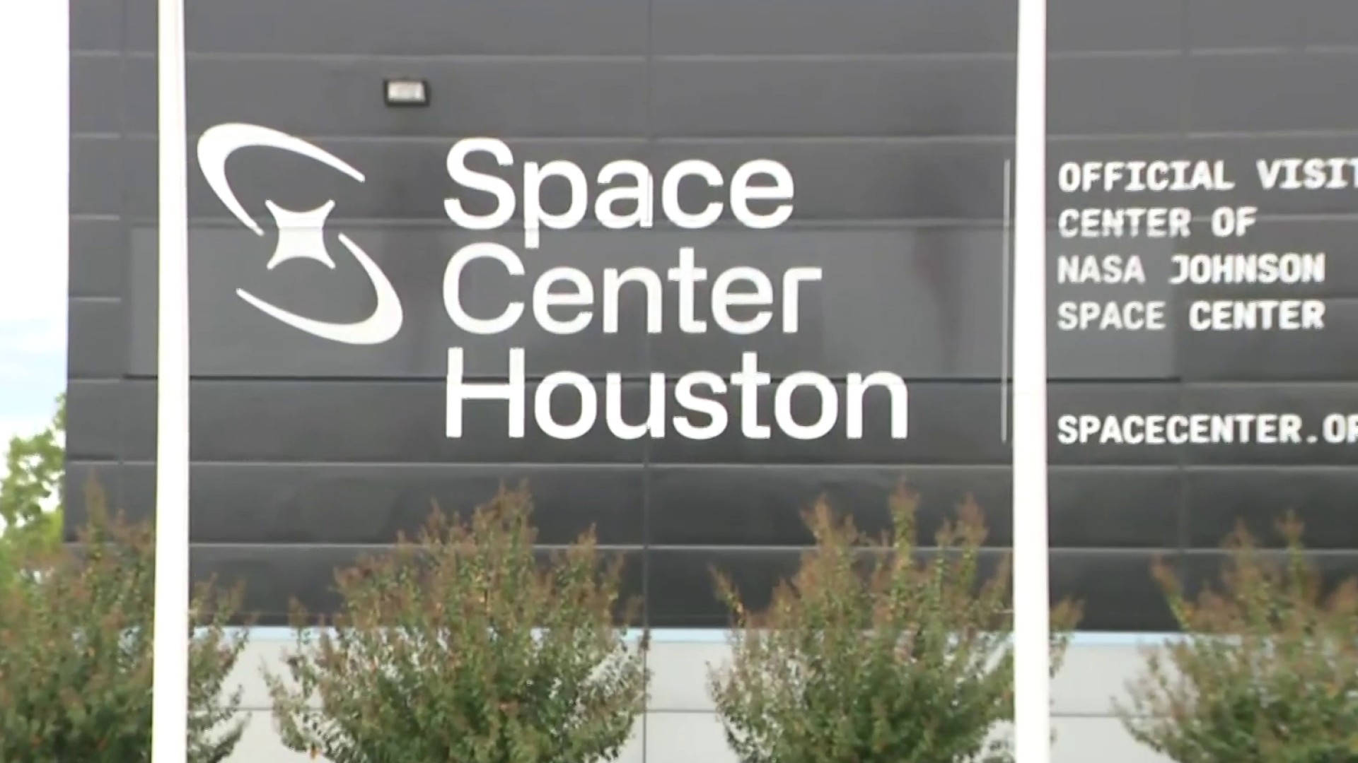 Logo 桌布 壁紙 — 現代黑色建築外牆展示白色「Space Center Houston」標誌，前景有綠色灌木。
