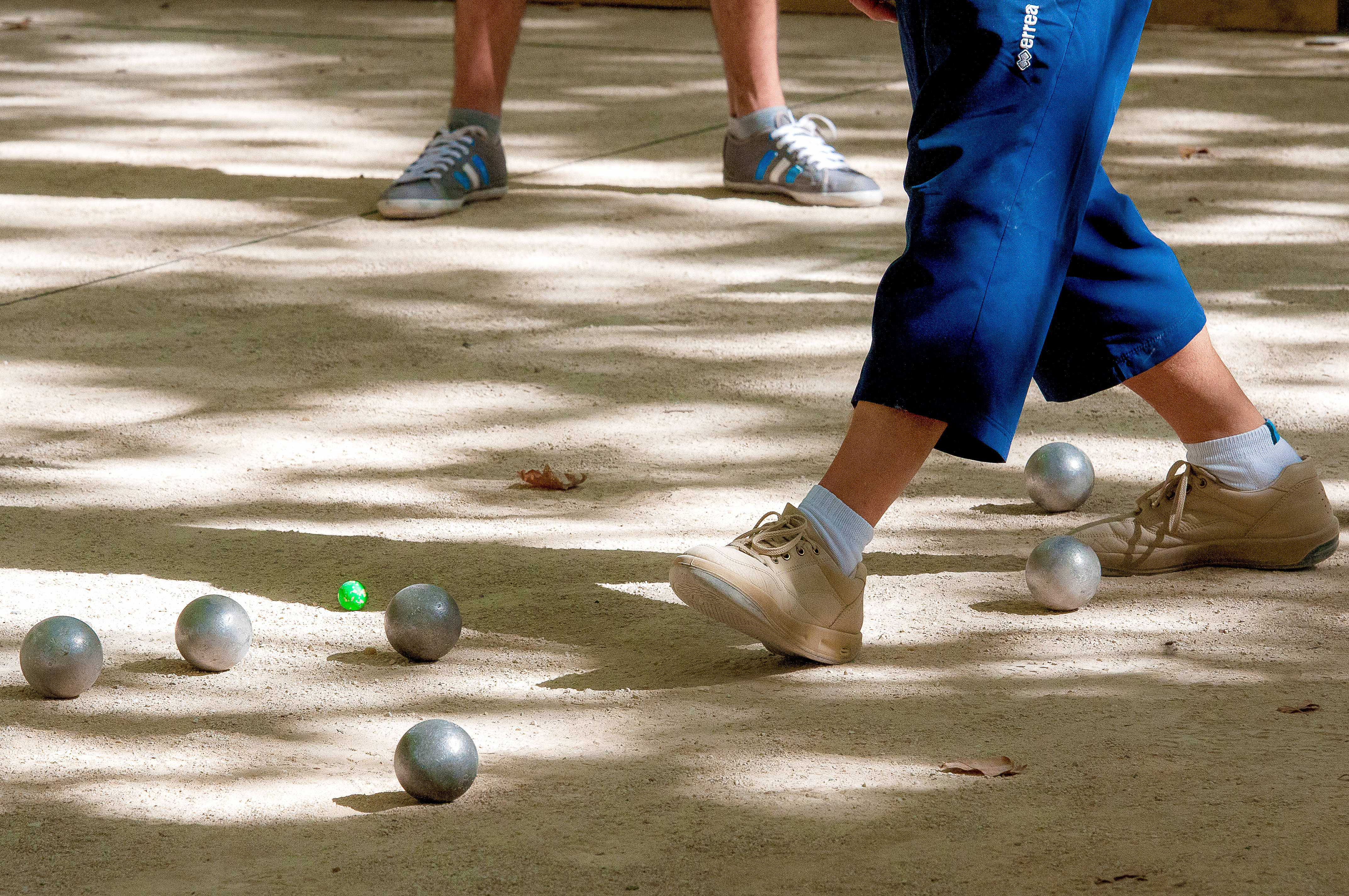 balls 桌布 壁紙 — 球員在陽光斑駁的混凝土球場上玩法式滾球時，腿部及腳部的特寫，銀色和一個綠色球散落各處。