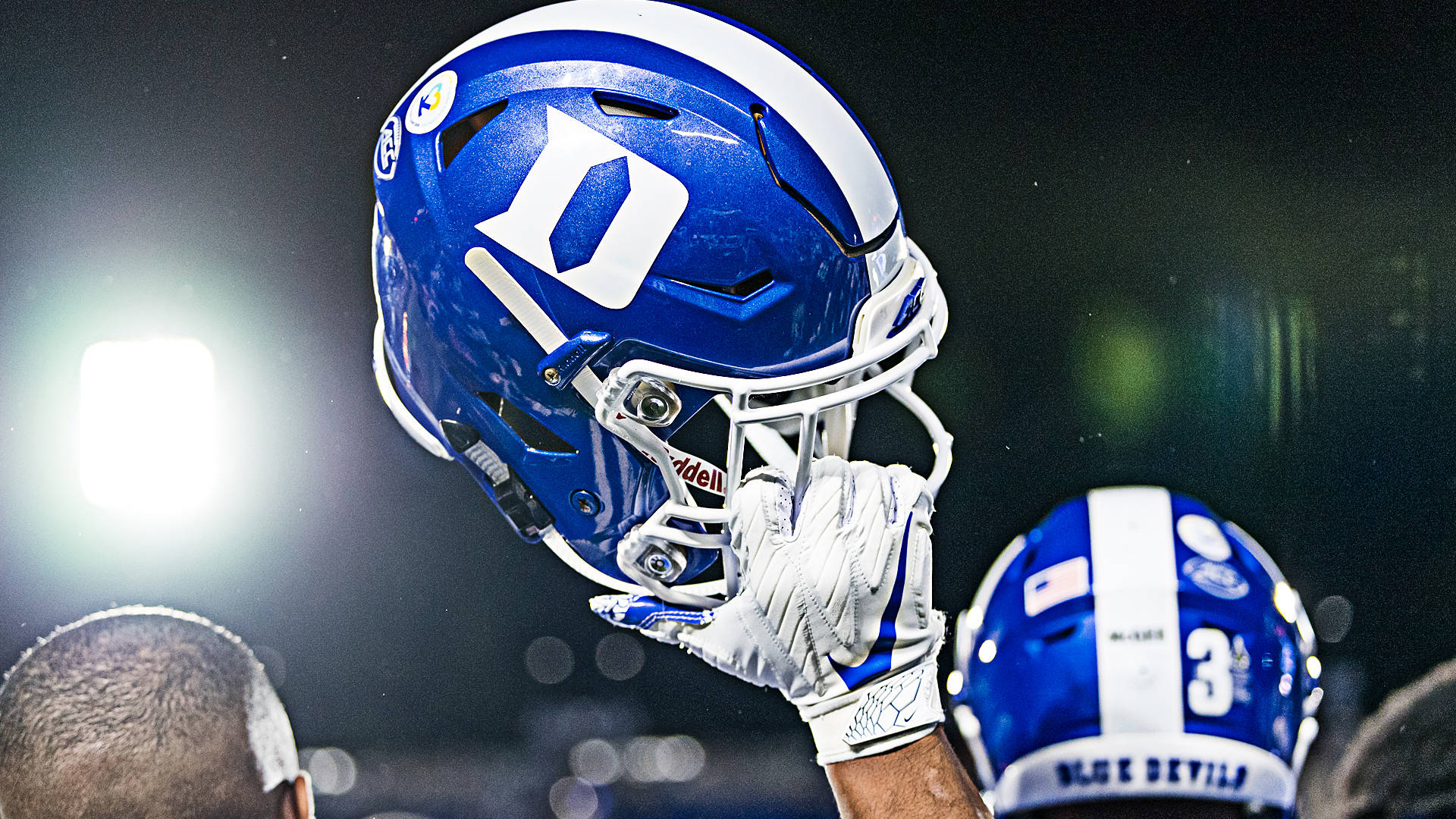 Duke Blue Devils 桌布 壁紙 — 球場燈光下，一名球員戴著手套的手高舉一個藍白相間的杜克大學美式足球頭盔的特寫。