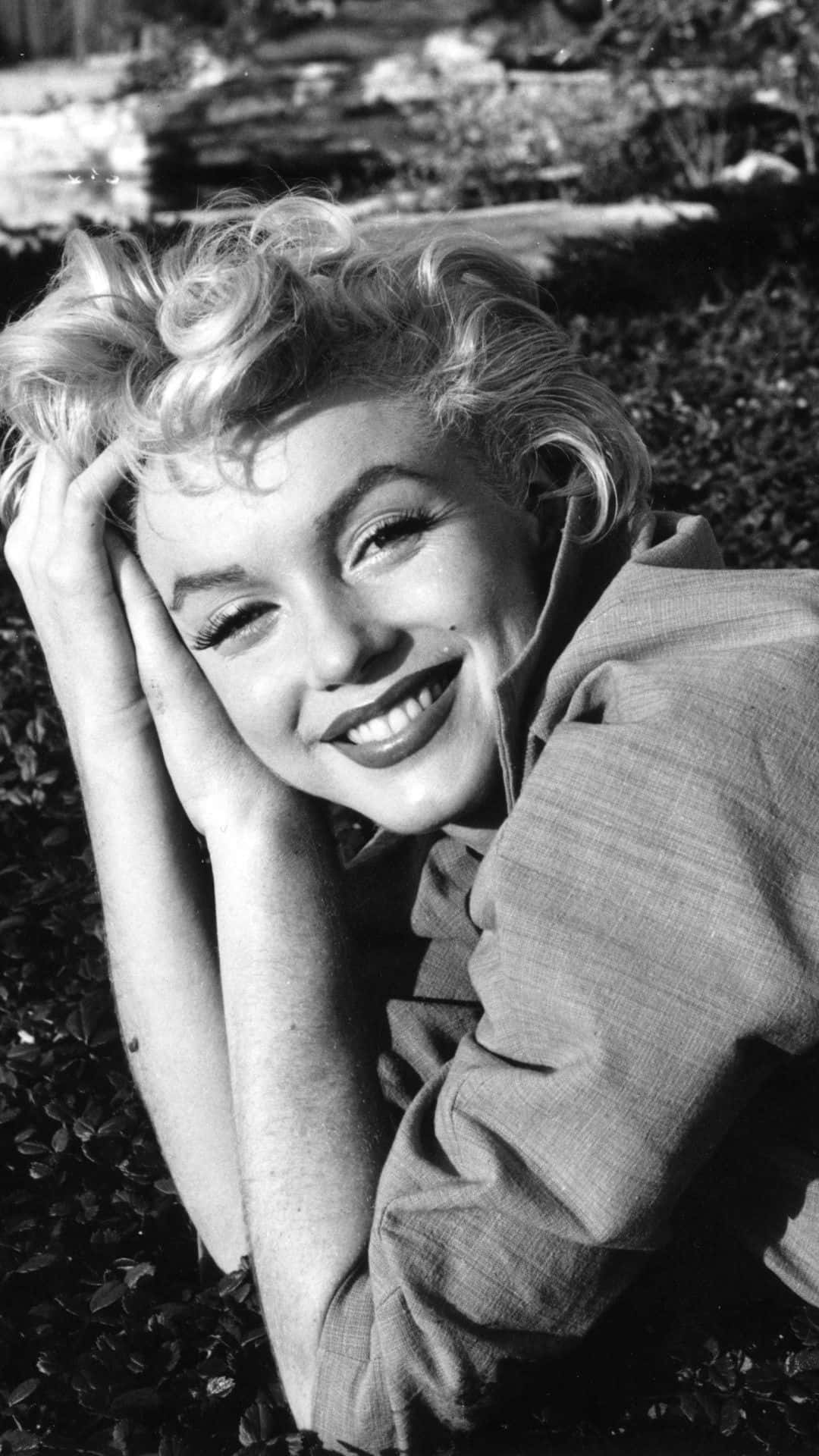 Marilyn Monroe iPhone 桌布 壁紙 — 瑪麗蓮夢露在草地戶外環境中將頭靠在手上，露出燦爛的笑容。