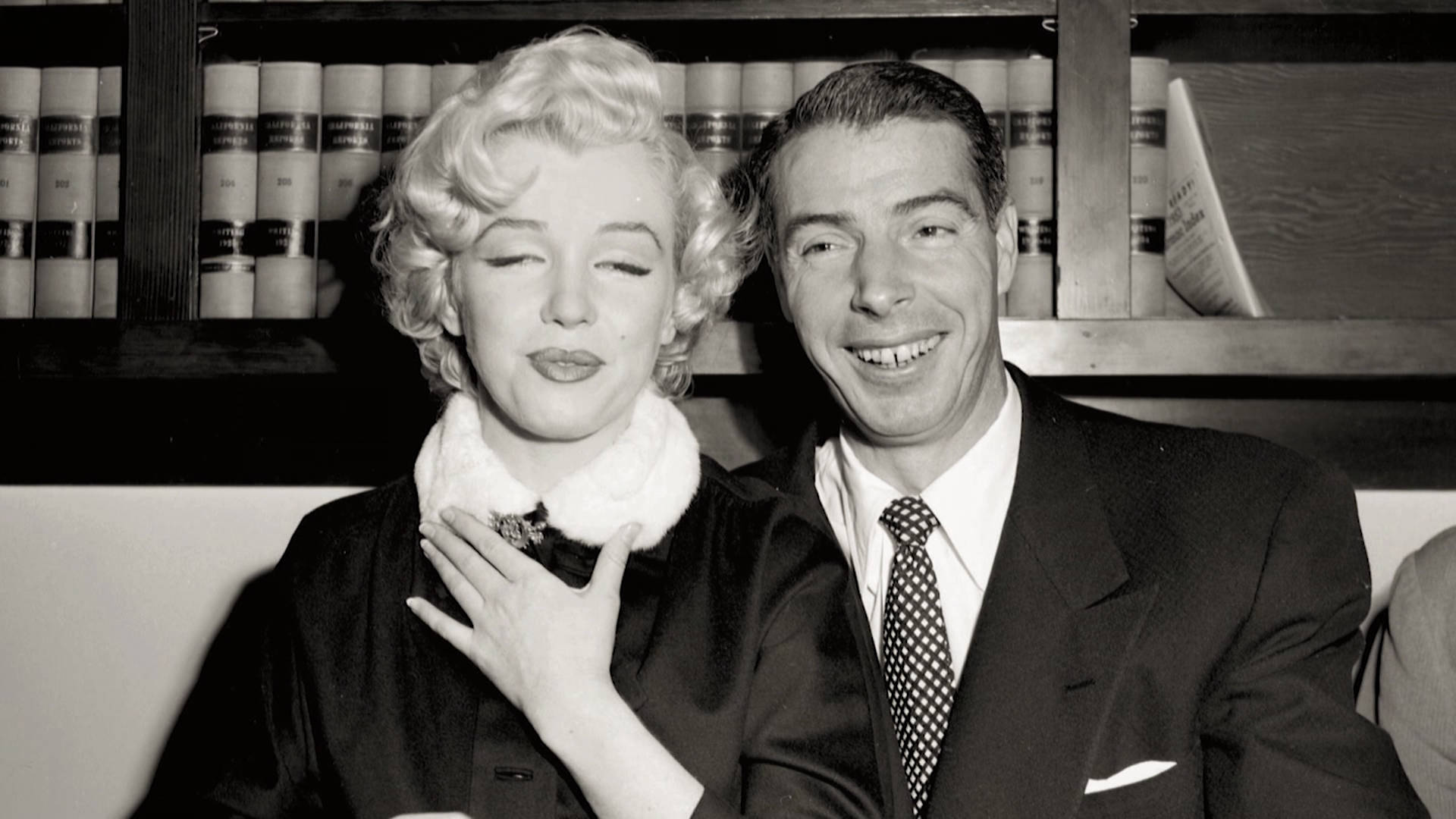 Joe Dimaggio 桌布 壁紙 — 瑪麗蓮夢露與一位微笑的男子在黑白照片中一起擺姿勢，身後有書架。