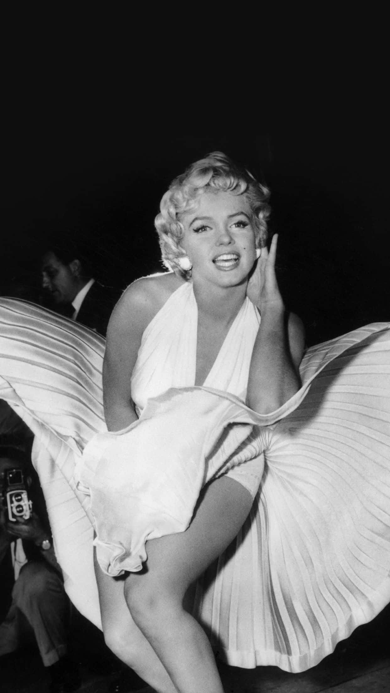 Marilyn Monroe iPhone 桌布 壁紙 — 瑪麗蓮夢露身穿白色百褶裙，在深色背景前將一隻手靠近臉部擺姿勢。