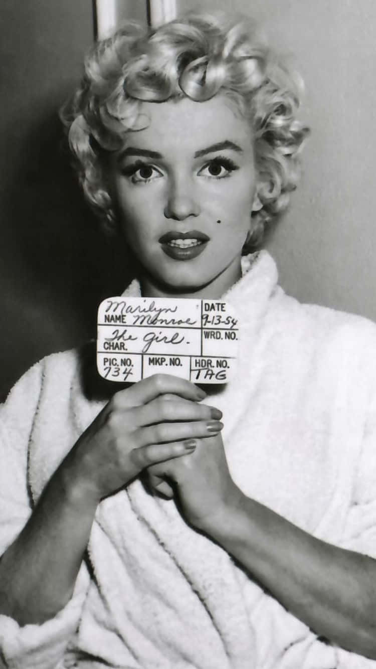 Marilyn Monroe iPhone 桌布 壁紙 — 瑪麗蓮夢露身穿白色長袍，手持一塊手寫的電影場記板，上面有她的名字和角色細節，這是一張經典的黑白工作室肖像照。