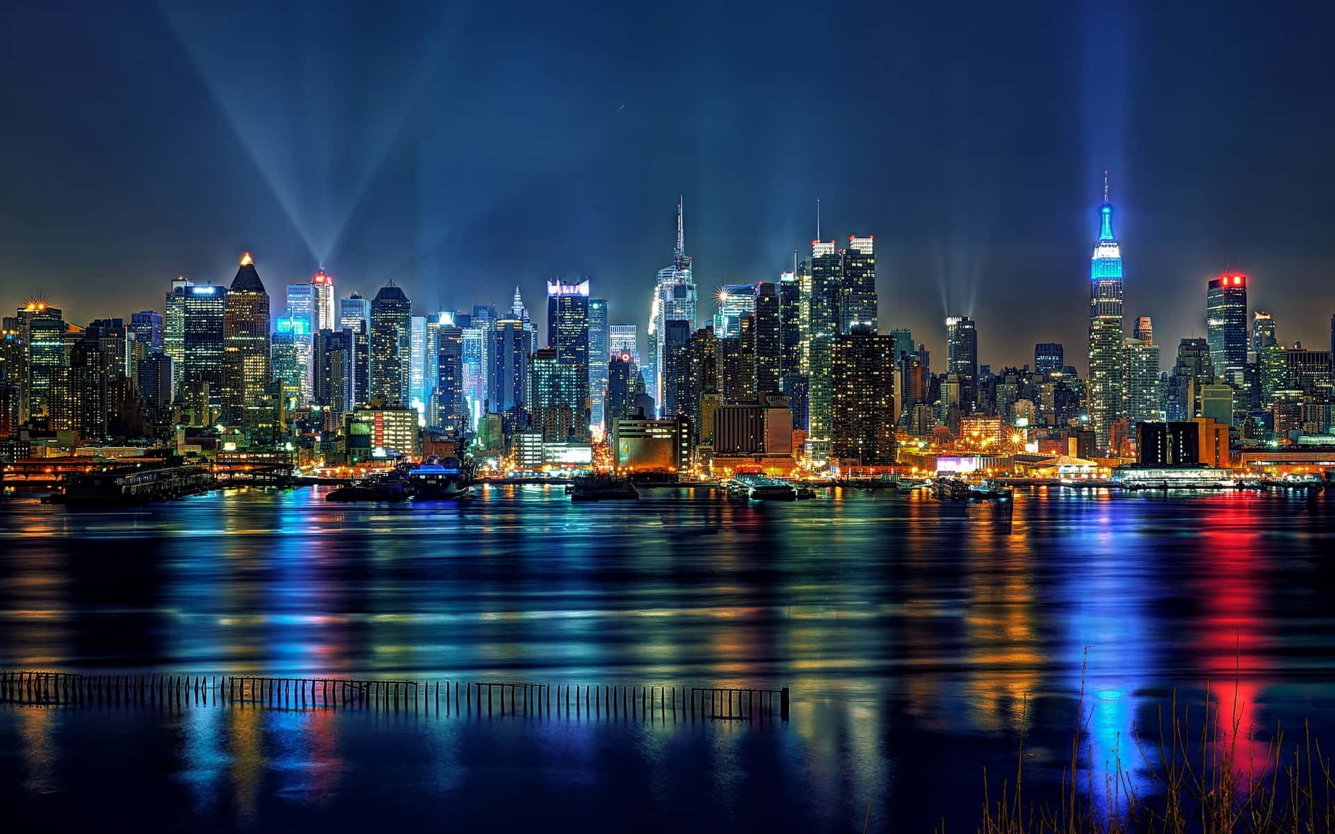 紐約市 桌布 壁紙 — 璀璨的夜景城市景觀，摩天大樓在黑暗的天空中閃耀，它們的燈光在平靜的水面上生動地反射。