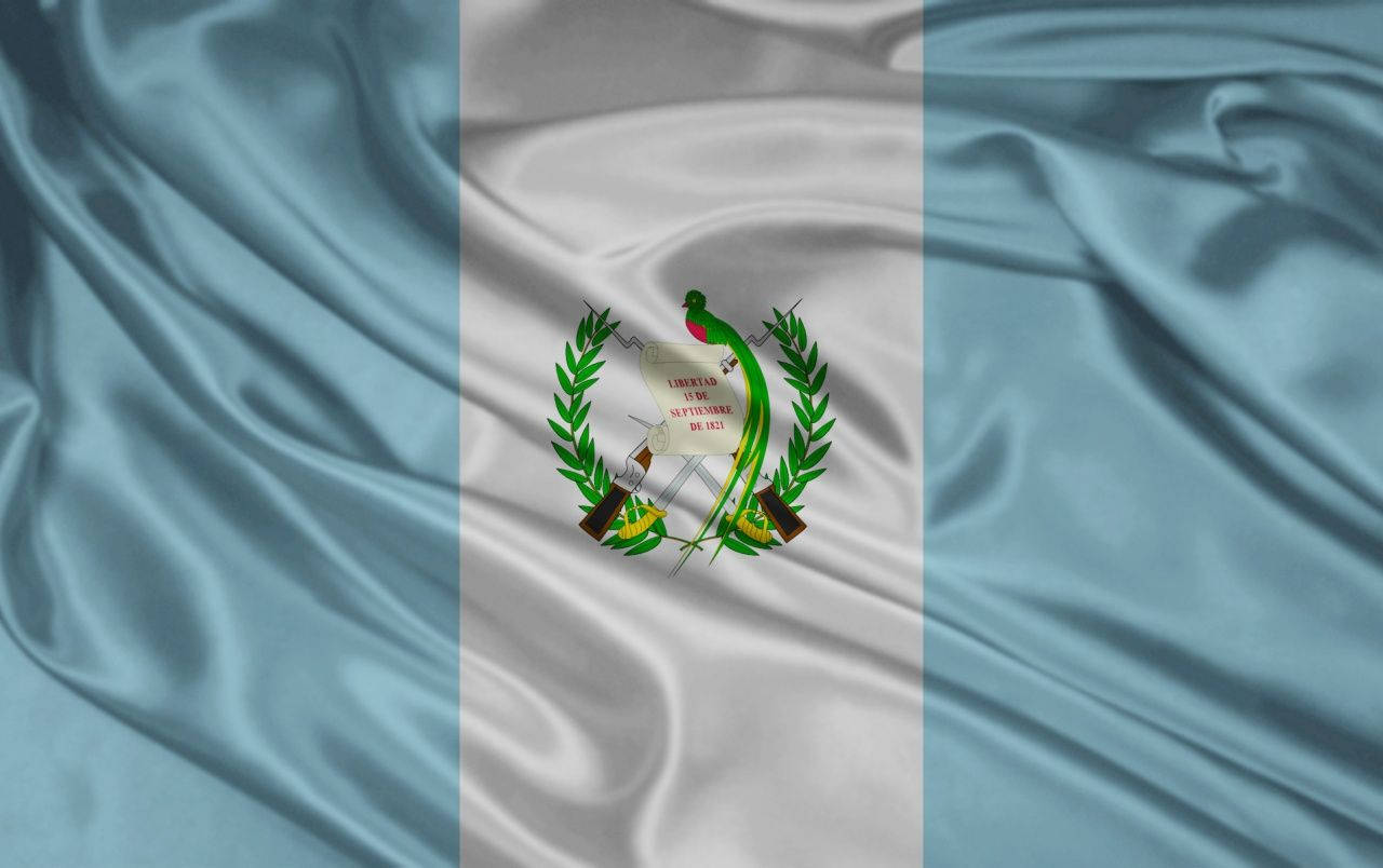 Guatemala 桌布 壁紙 — 瓜地馬拉的國旗在柔軟的布料褶皺中飄揚，帶有淺藍色和白色條紋，中央有包含格查爾鳥的盾徽。