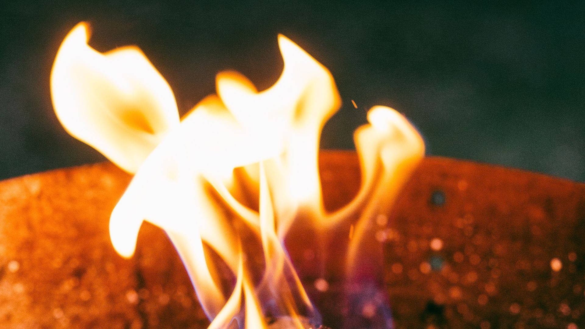 Flame 桌布 壁紙 — 生動的特寫鏡頭，捕捉到從生鏽金屬火坑中升起的舞動橙色和黃色火焰，背景為深色。