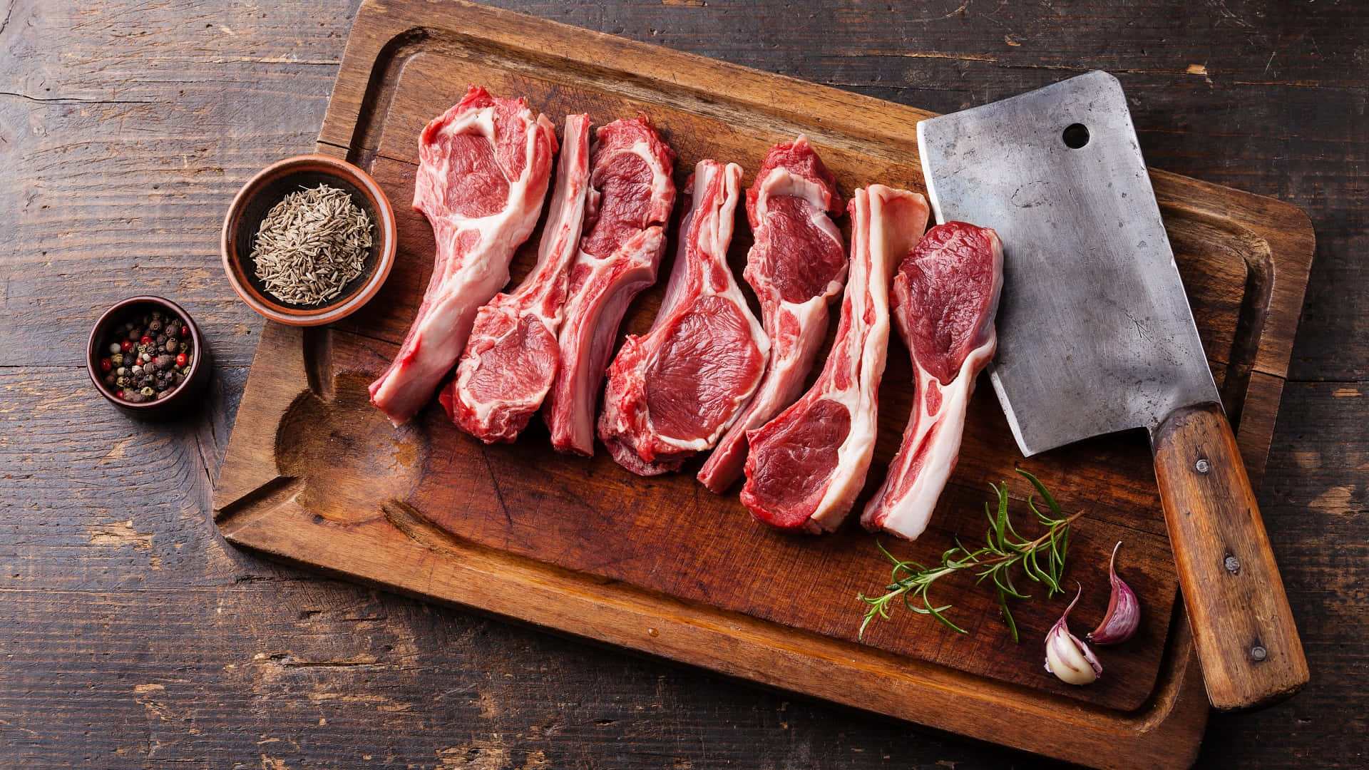 meat 桌布 壁紙 — 生羊排搭配迷迭香、大蒜和香料，擺放在質樸的木砧板上，附一把剁肉刀。