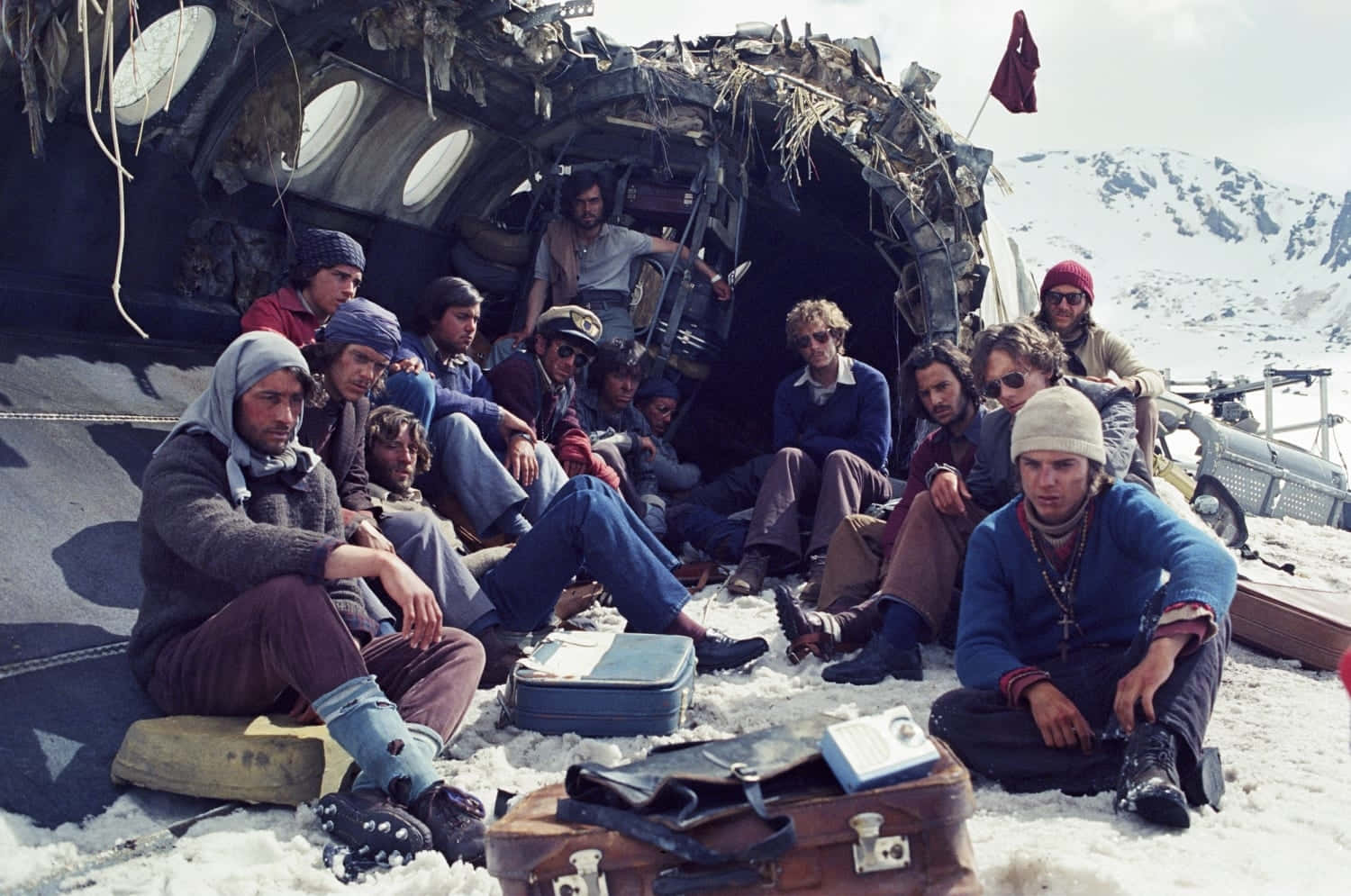 the society 桌布 壁紙 — 生還者坐在雪地上，旁邊是失事飛機的機身，背景是山區冬季景觀。