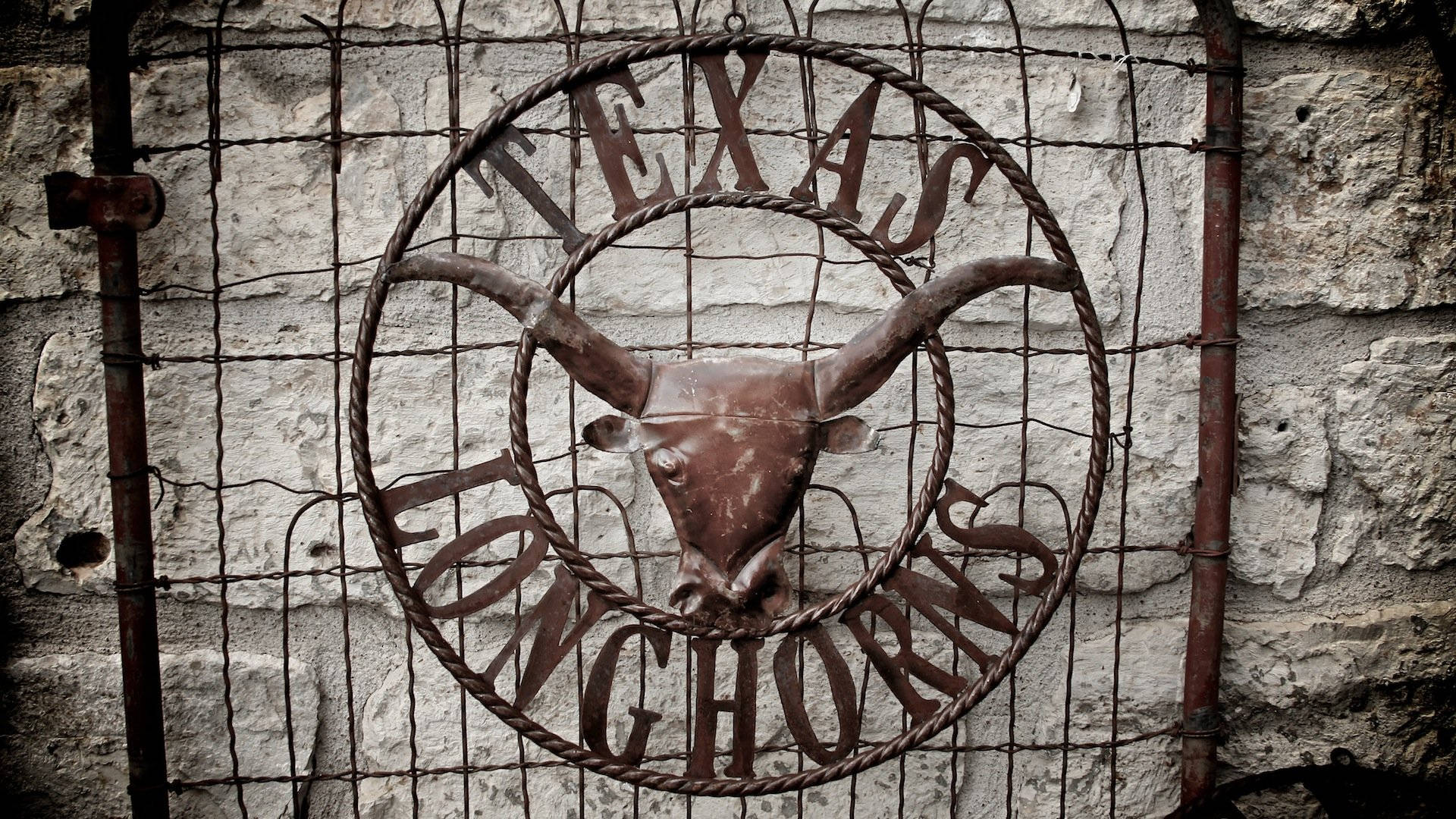 桌布 壁紙 — 生鏽的金屬長角牛標誌，上面有「TEXAS LONGHORNS」字樣，掛在有刺鐵絲網圍欄後面的風化石牆上。