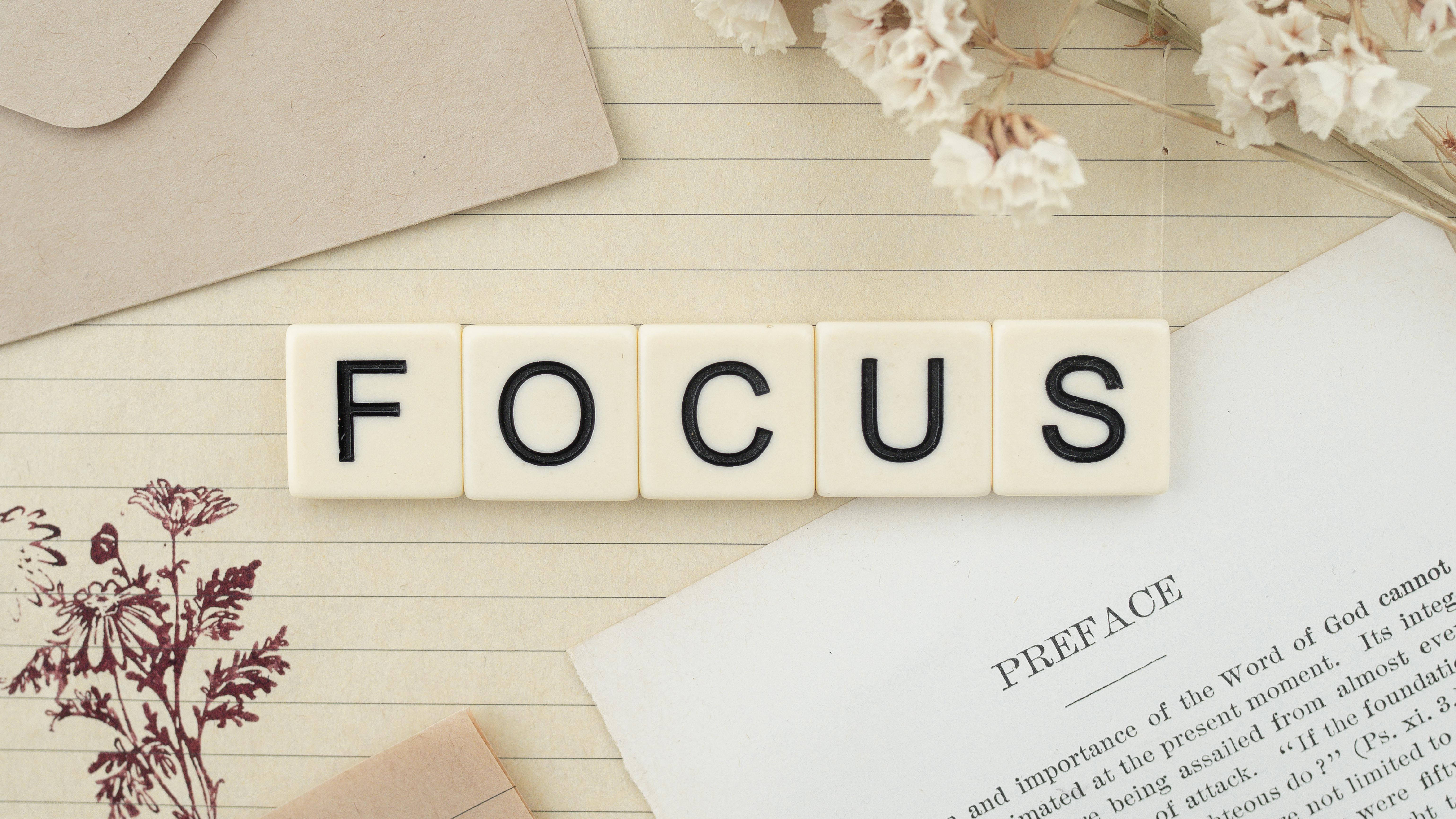 勵志動圖 桌布 壁紙 — 用奶油色瓷磚拼出的單詞FOCUS放在橫線紙上，旁邊有乾燥花和一本打開的書。