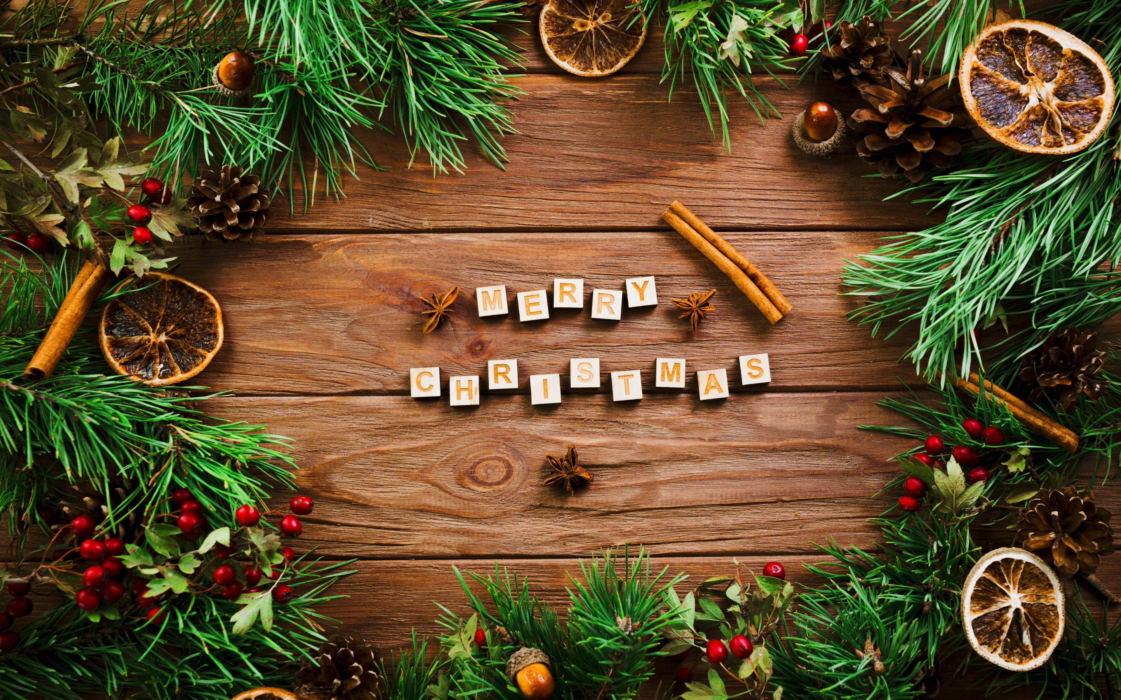 美麗聖誕 桌布 壁紙 — 用木製字母拼出的「聖誕快樂」，周圍環繞著松樹枝、肉桂棒和乾橙，放在一張質樸的木桌上。