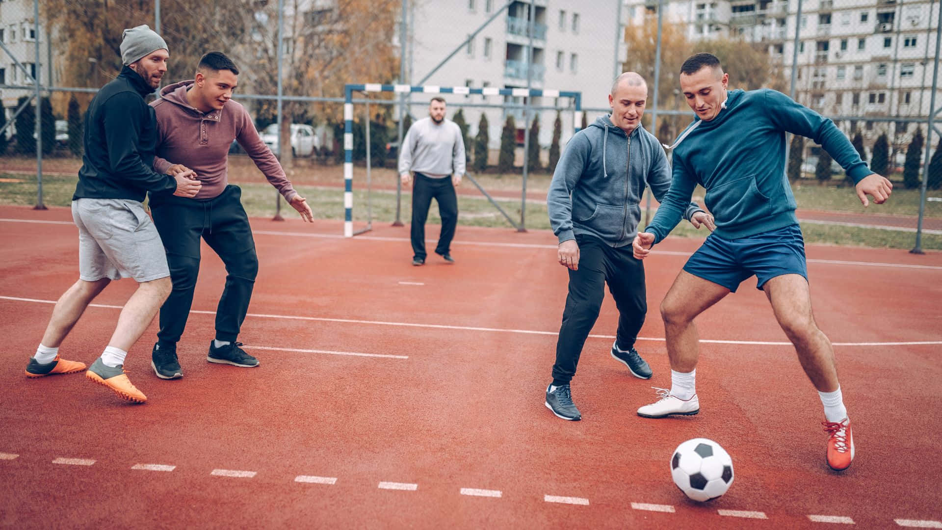 Best Sports 桌布 壁紙 — 男人在紅色球場上踢足球，背景有秋天的樹木和公寓大樓。